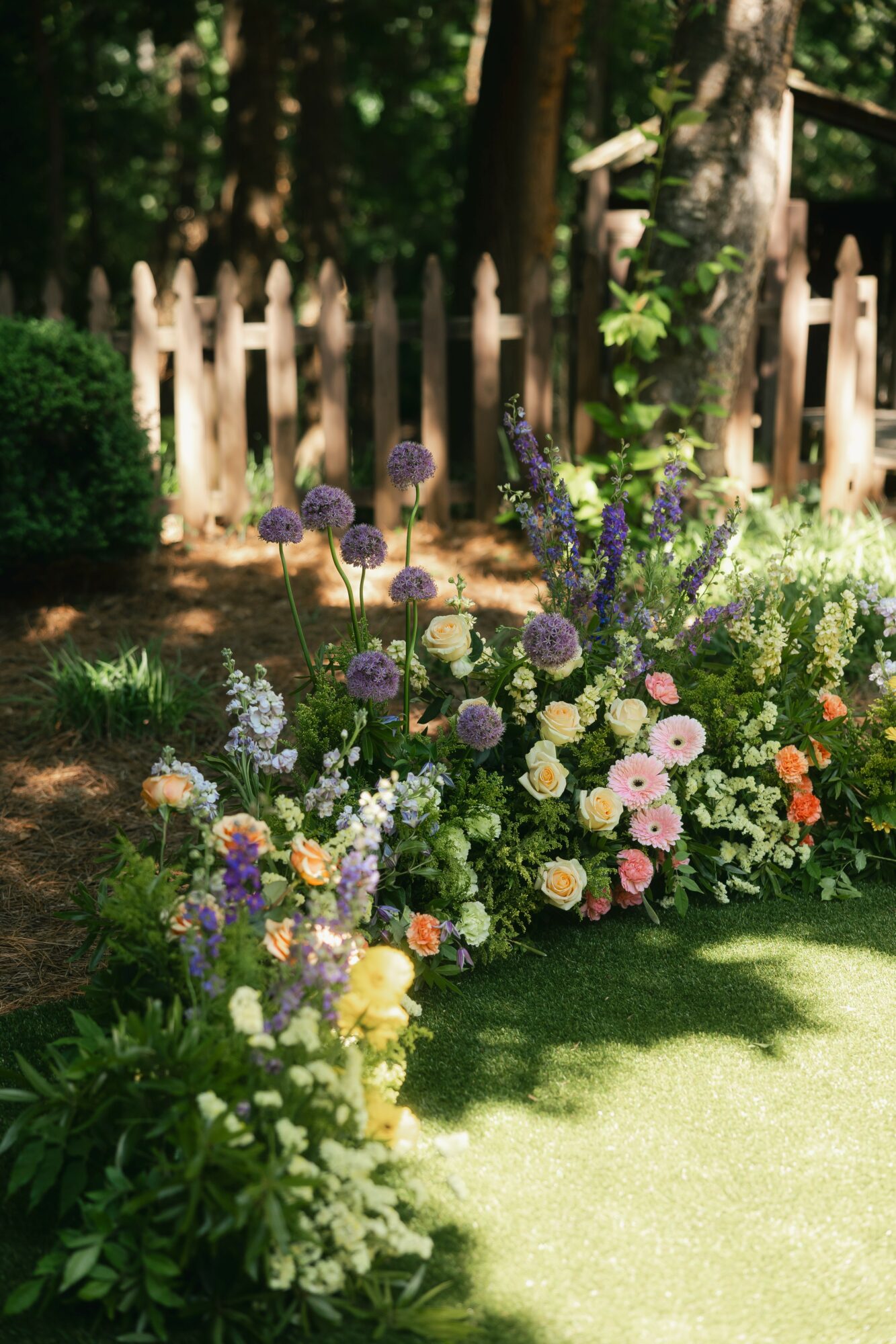 Colorful flower bed with purple, pink, white, and orange flowers in front of a wooden fence and trees, sunlight shining.