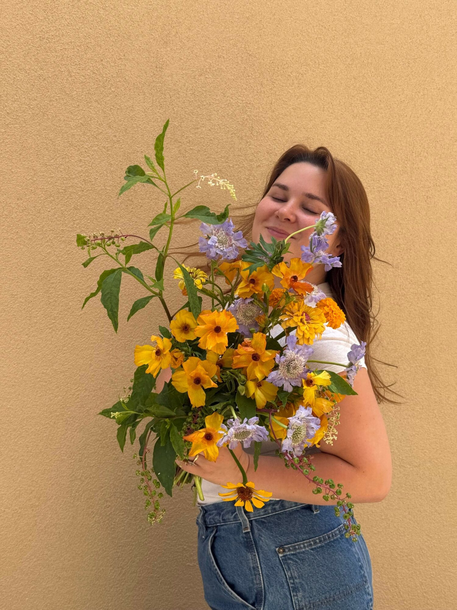 Woman holding a large bouquet of yellow, purple, and white flowers, standing against a beige wall.