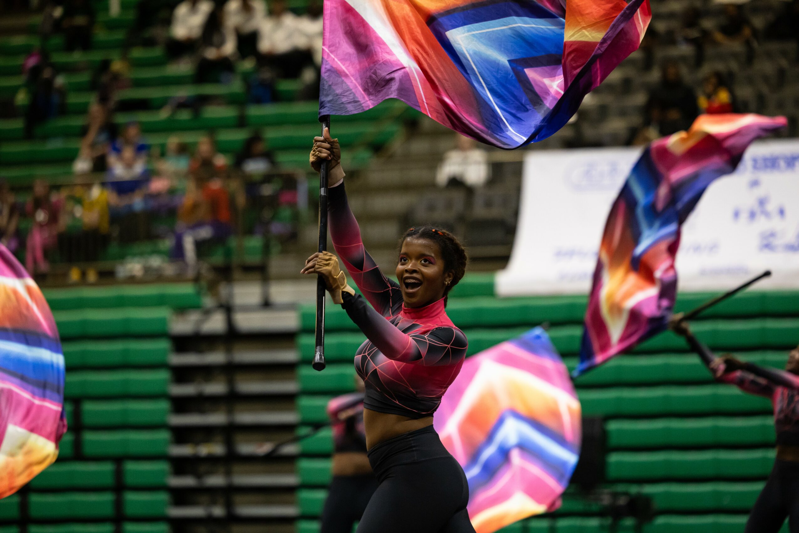 Person smiling and waving colorful flags in a stadium with spectators in the background.