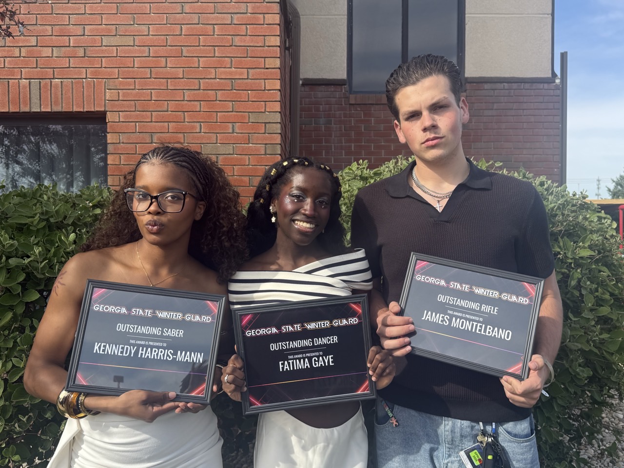Three young people holding award plaques outdoors in front of a brick building and greenery, smiling at the camera.