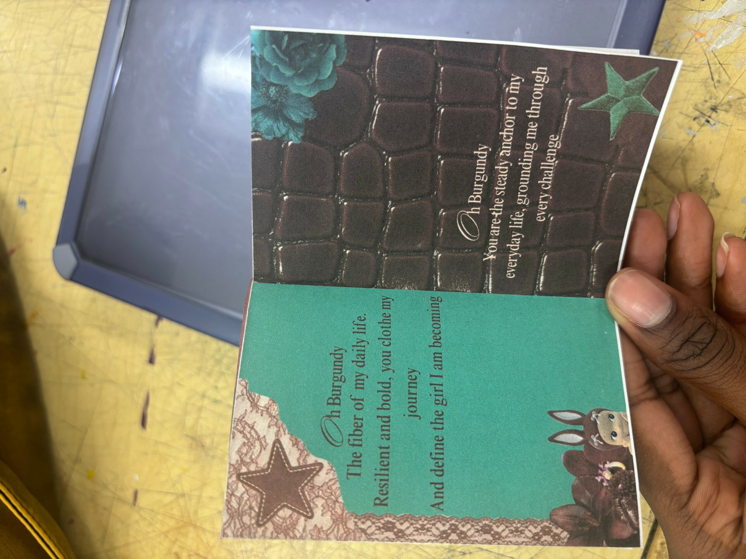 Hand holding a card with a star, bunny, and text, against a yellow surface and blue box.