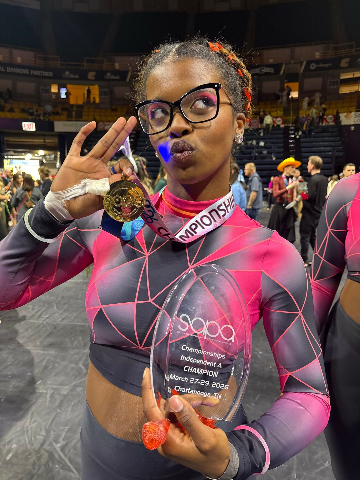 Woman in athletic clothing holding a medal and a glass award, wearing glasses and a pink and black sports outfit.