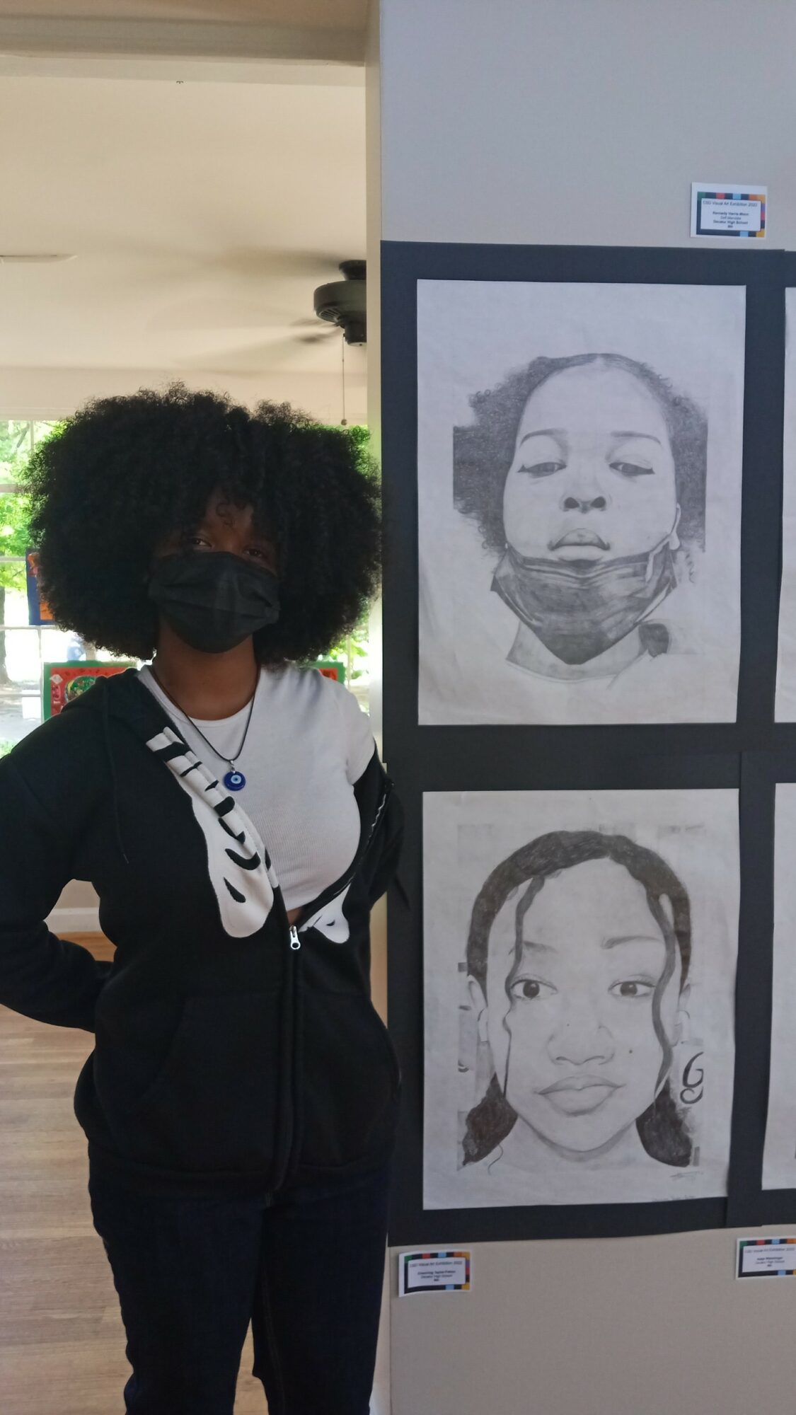 Person with curly hair, wearing a face mask and black jacket, standing next to black-and-white portrait drawings on a wall.