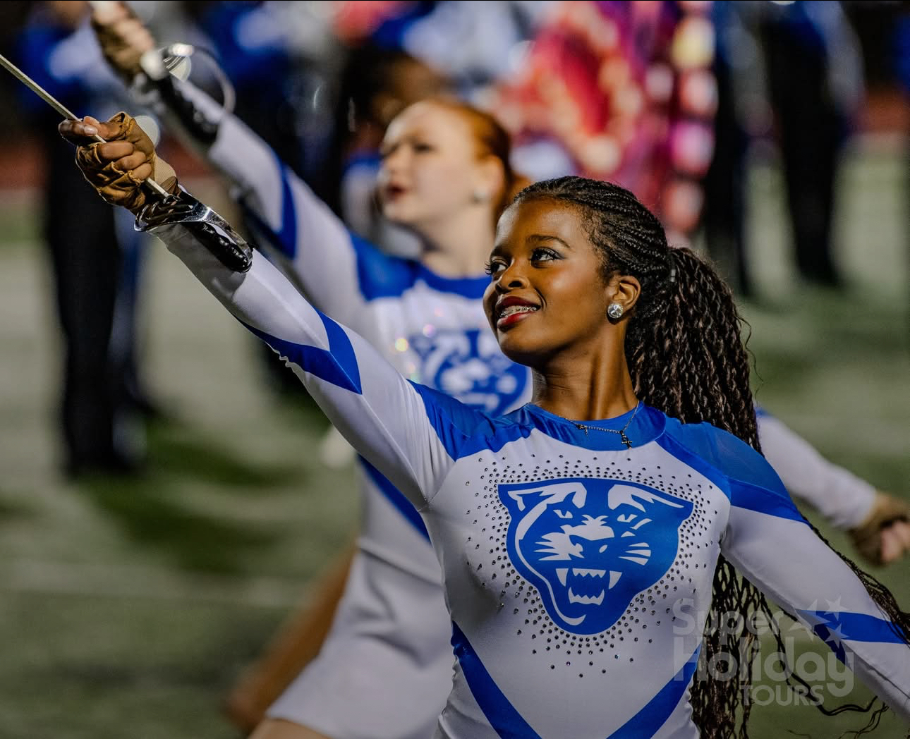 Cheerleader with braided hair in a blue and white uniform, raising her arm during a performance, with other cheerleaders in background.