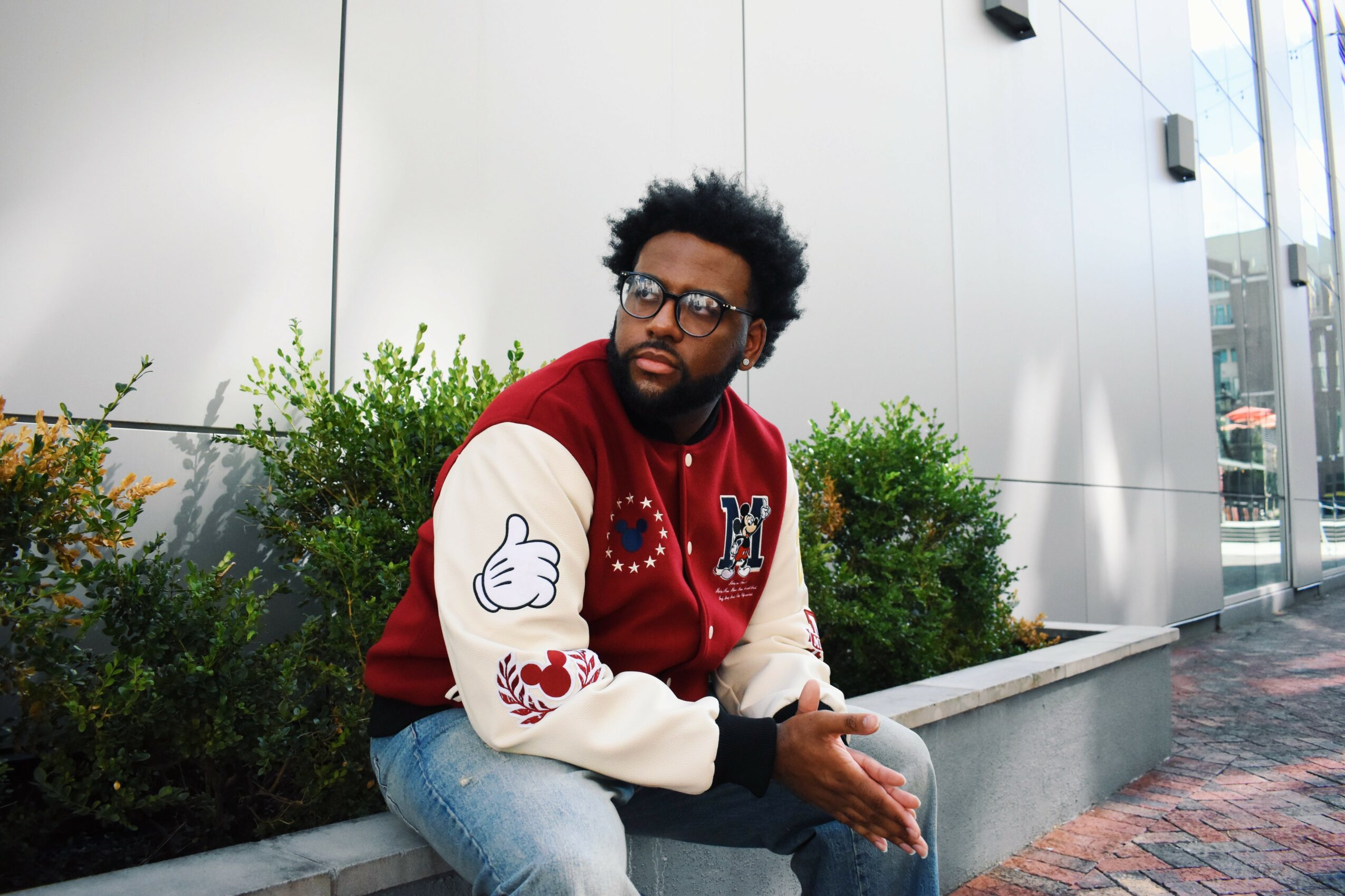 Man with glasses and beard sitting outdoors near plants, wearing a red and white varsity jacket and jeans.
