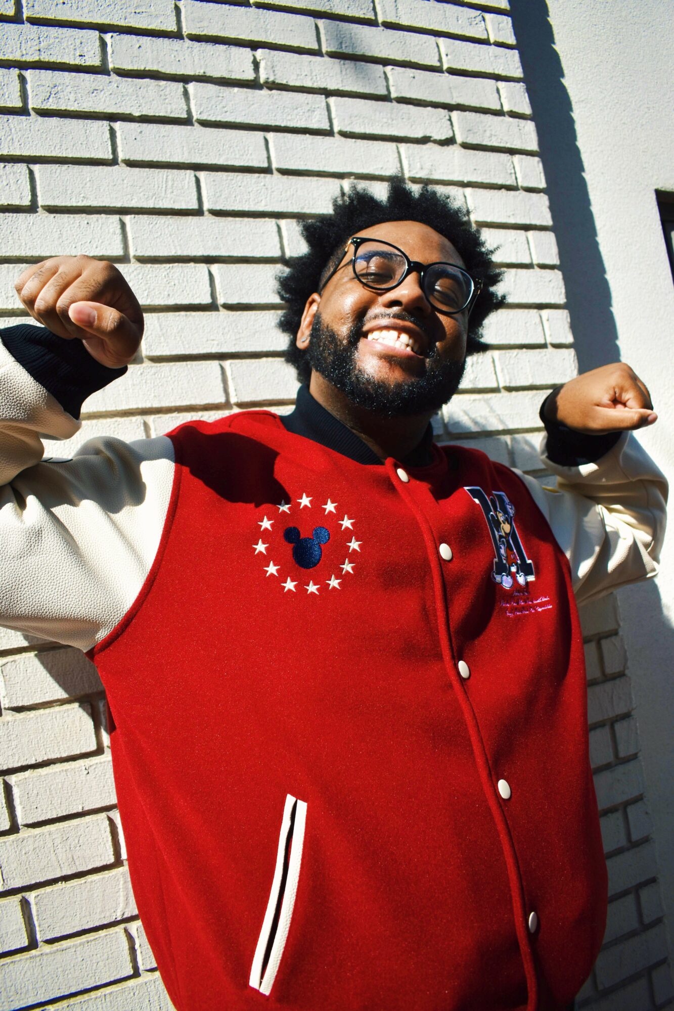 Person smiling and flexing arms outdoors against a brick wall, wearing glasses and a red jacket with patches.