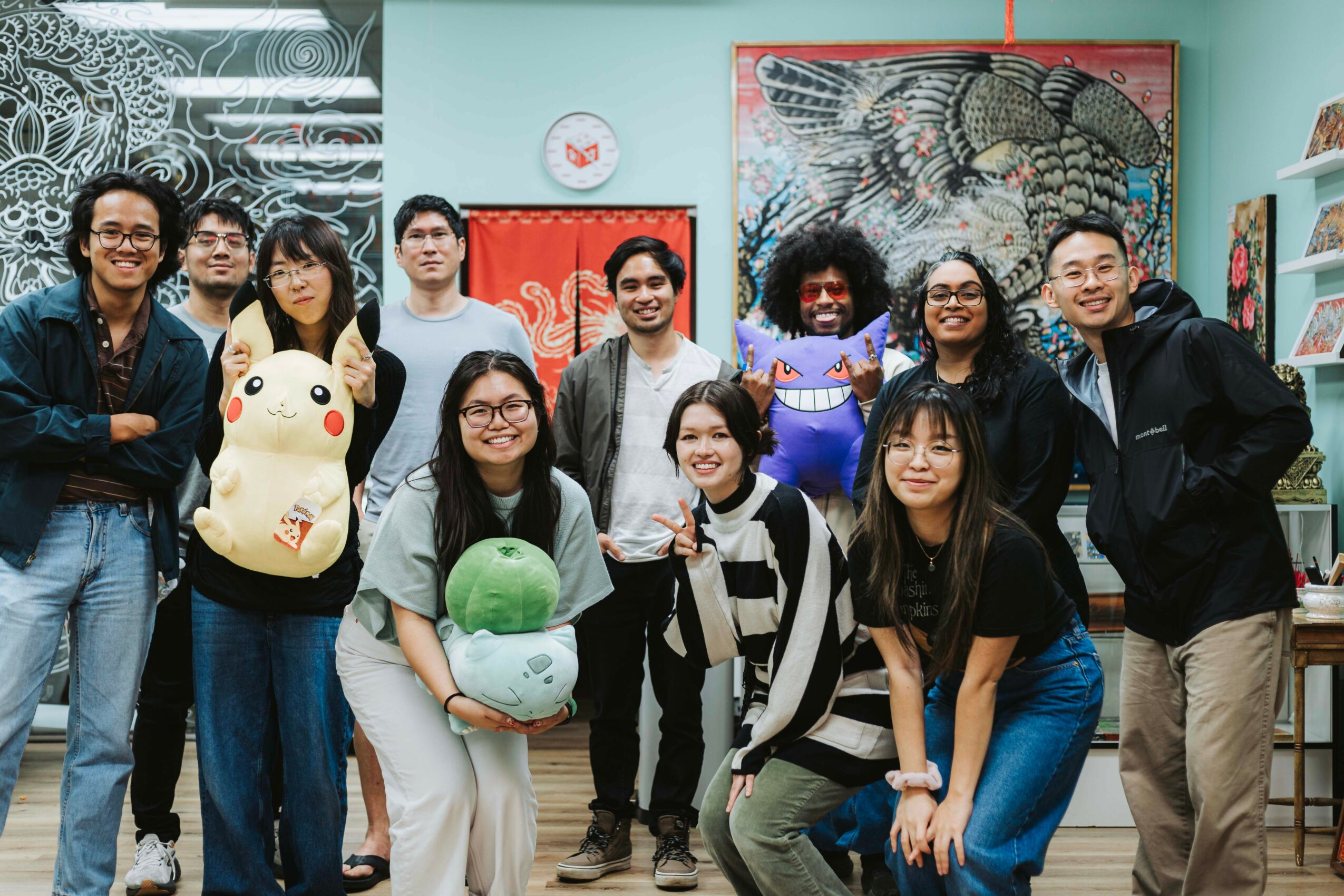 Group of ten people smiling indoors, holding plush toys, with colorful artwork on the wall behind them.