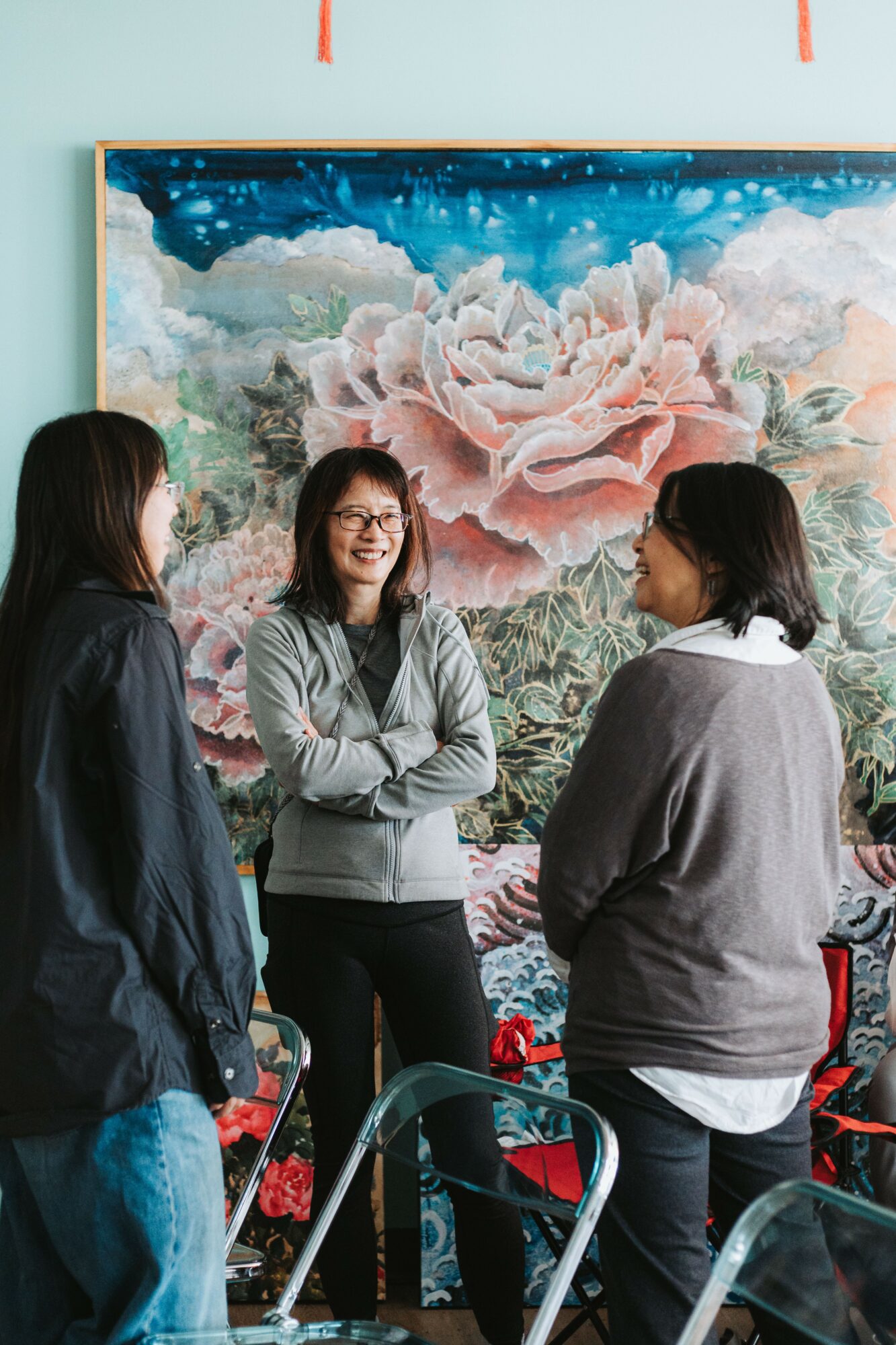 Three women standing and talking in front of a large floral painting, smiling and engaging in conversation.