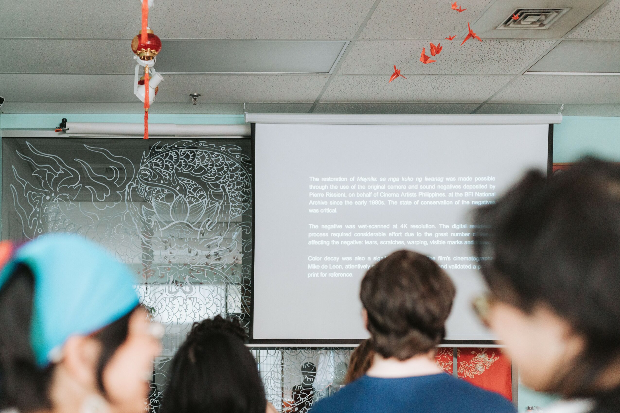 People watching a presentation in a room with a projection screen and decorative wall, some wearing glasses and head coverings.
