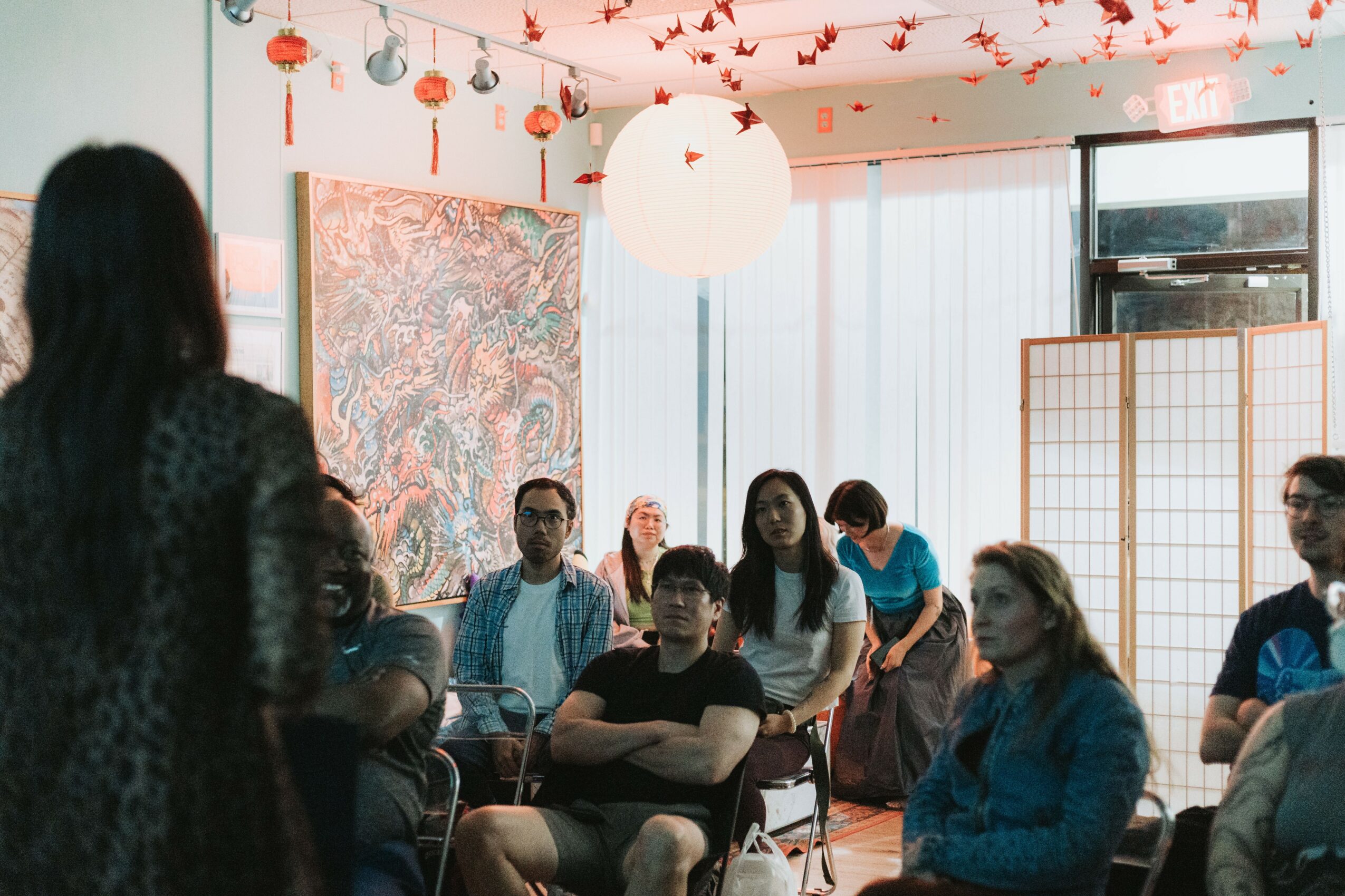 Group of people seated indoors, listening to a person speaking, with colorful decorations and large windows in the background.