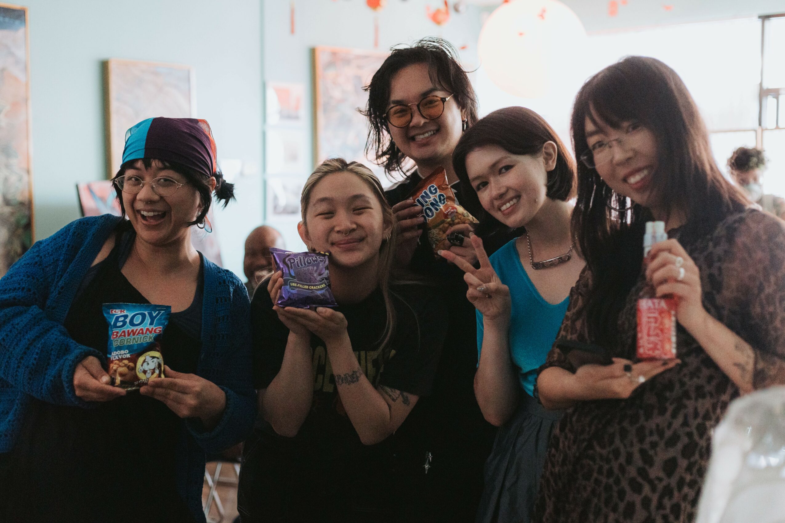 Group of five women smiling and holding snacks indoors, some wearing glasses, in a well-lit room with artwork on the walls.