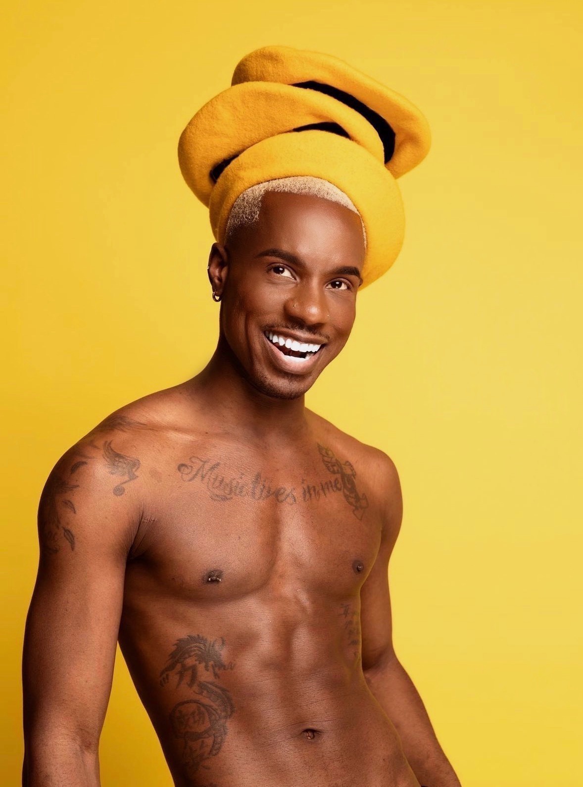 Smiling shirtless man with tattoos and a yellow headwrap against a yellow background.
