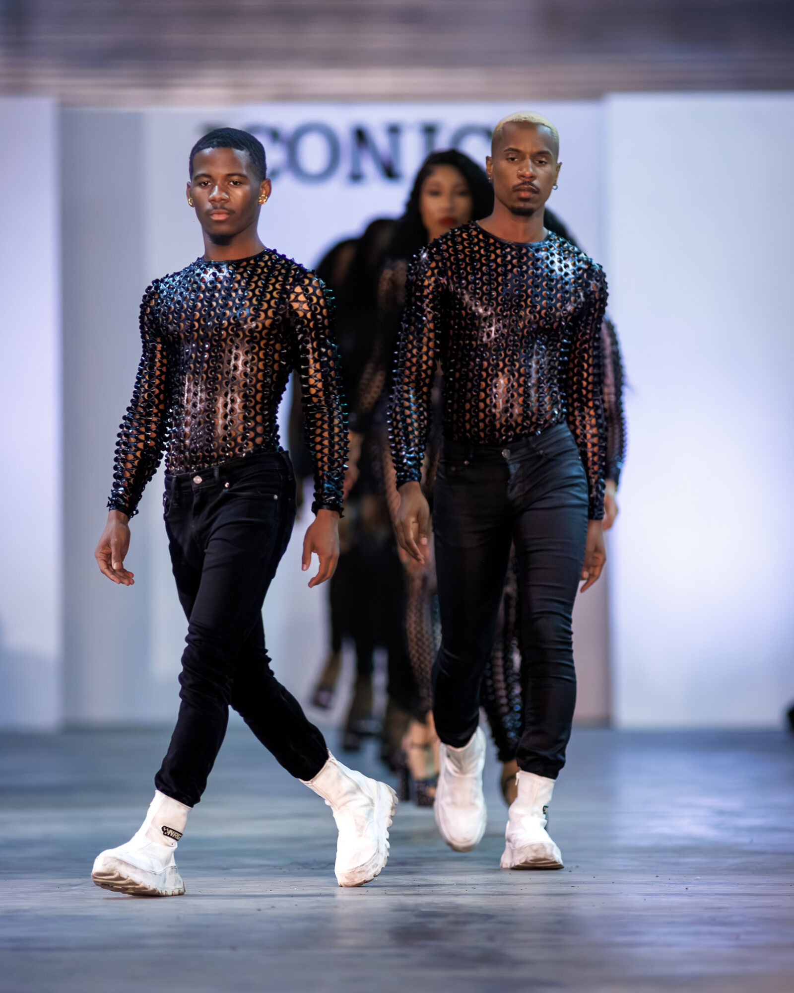 Three models walk down a runway wearing black outfits with shiny, perforated tops and white sneakers.