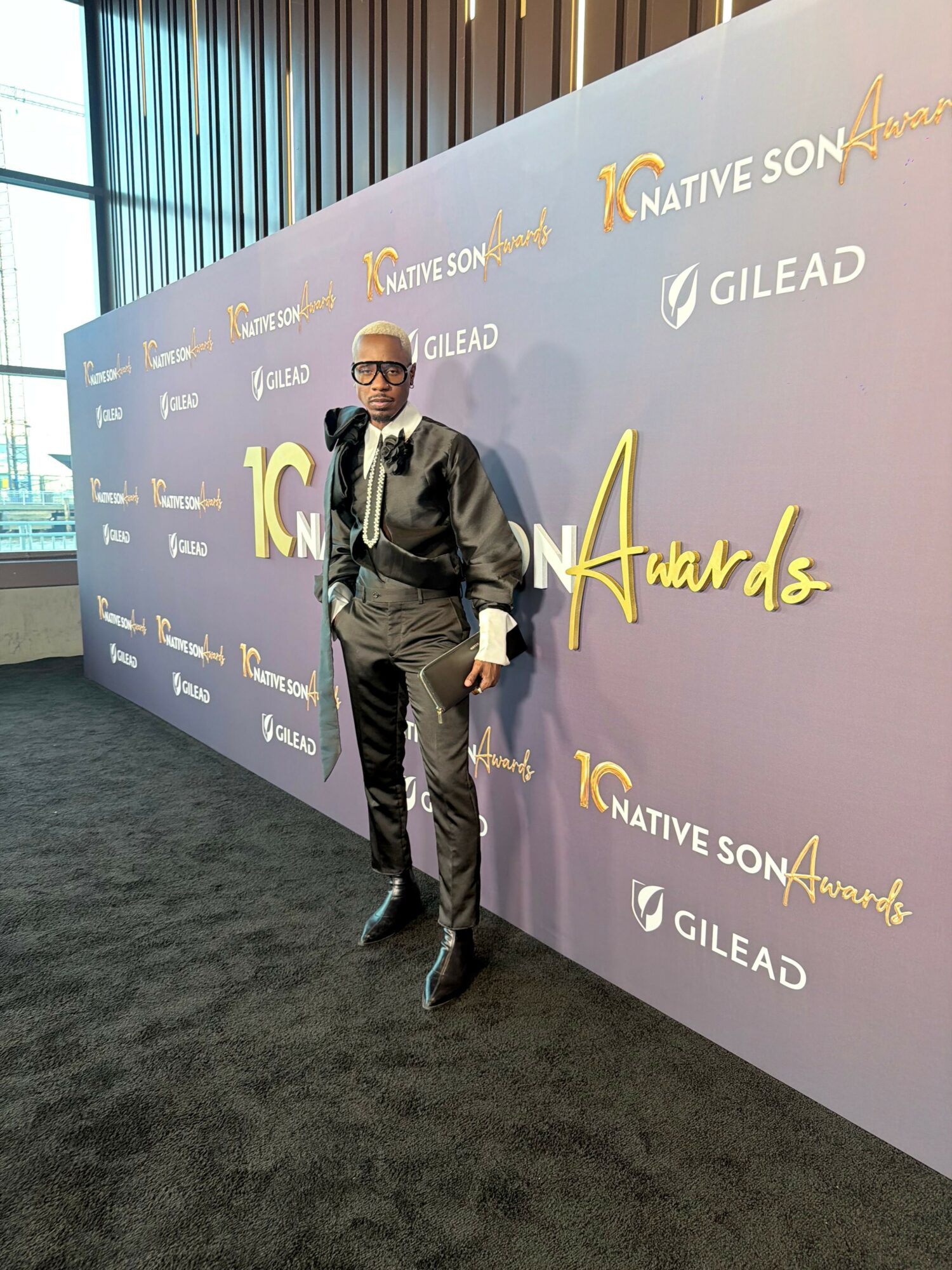 Person in black jacket and pants standing on black carpet in front of a purple backdrop with gold and white text.