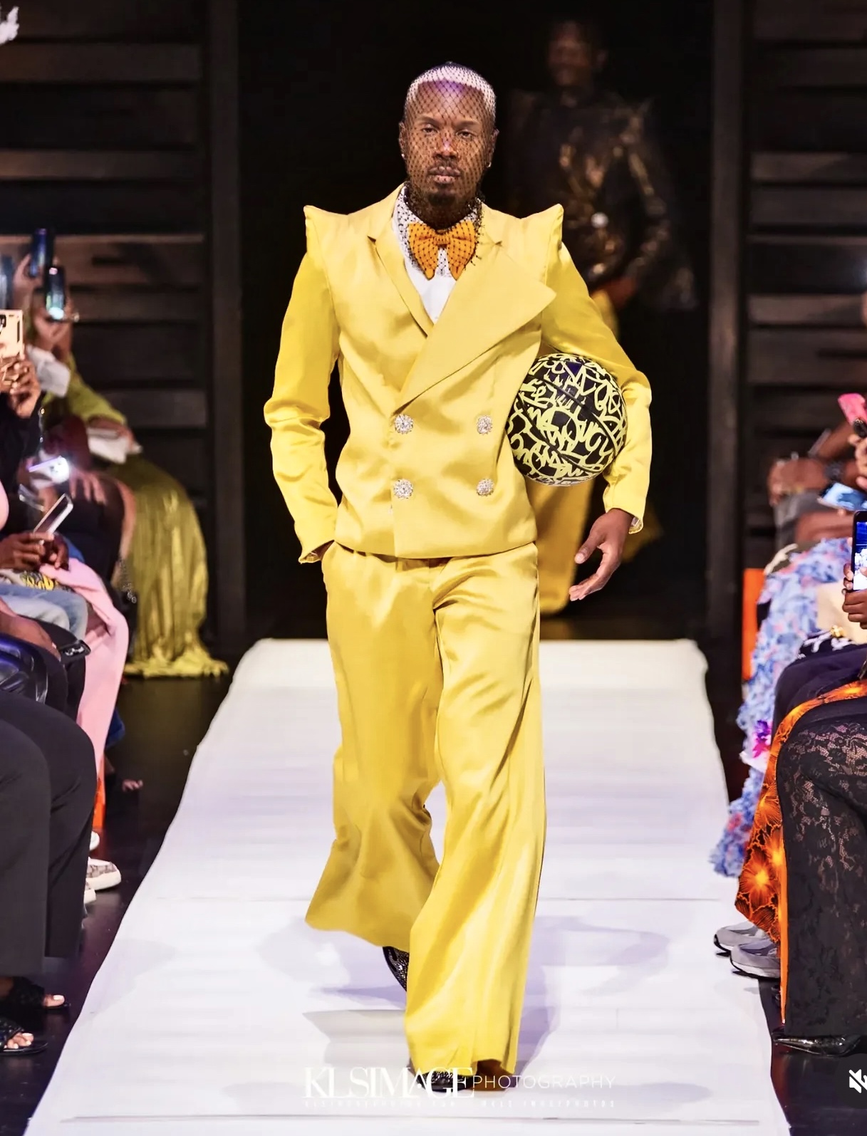 Male model walking down runway in bright yellow suit holding a patterned hat, audience seated on both sides, dark background
