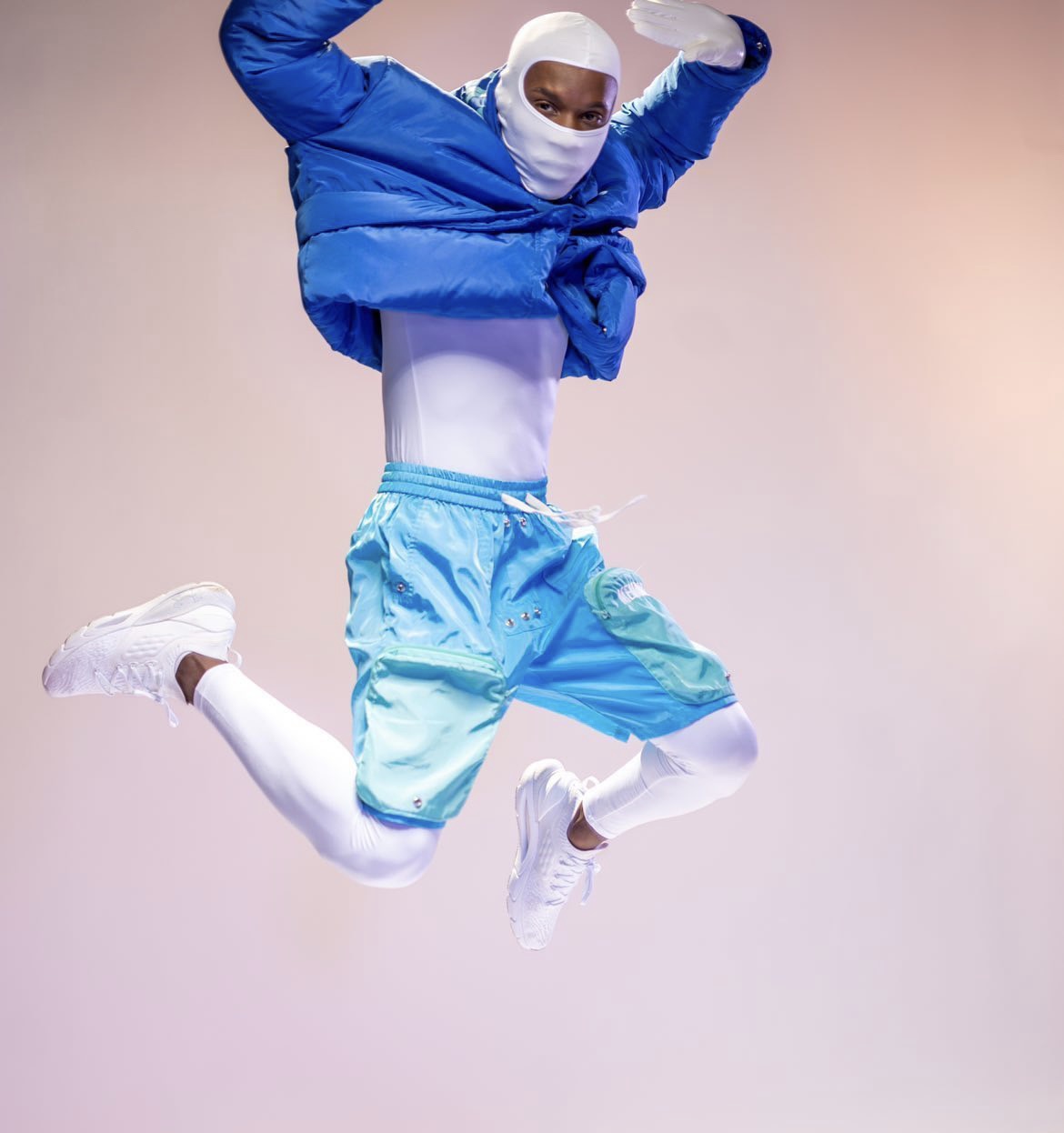 Person jumping mid-air wearing a blue jacket, light blue shorts, white leggings, and white sneakers, with a white head covering and face mask.
