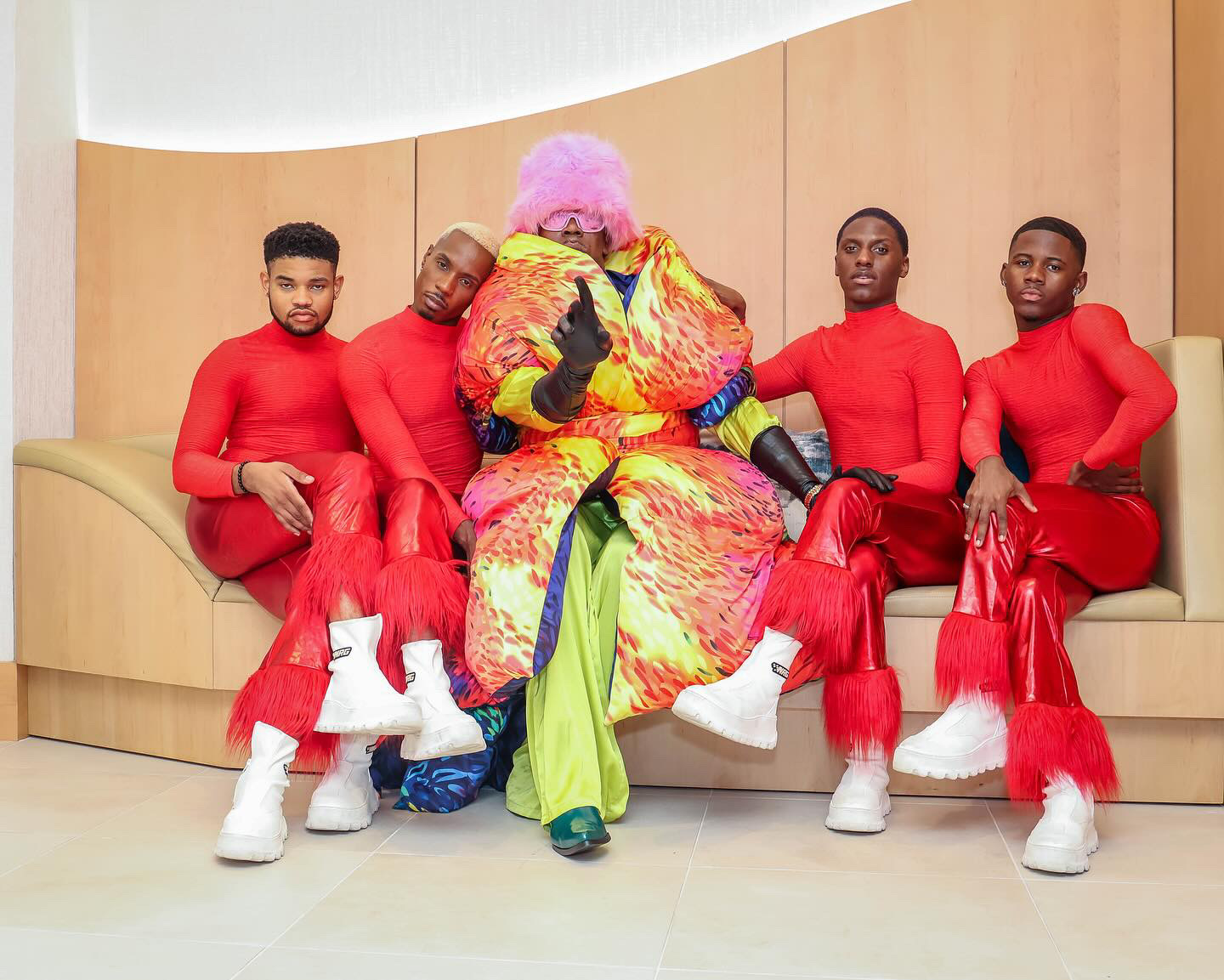 Person in colorful costume with pink hair sitting with four men in red outfits on a curved beige bench.