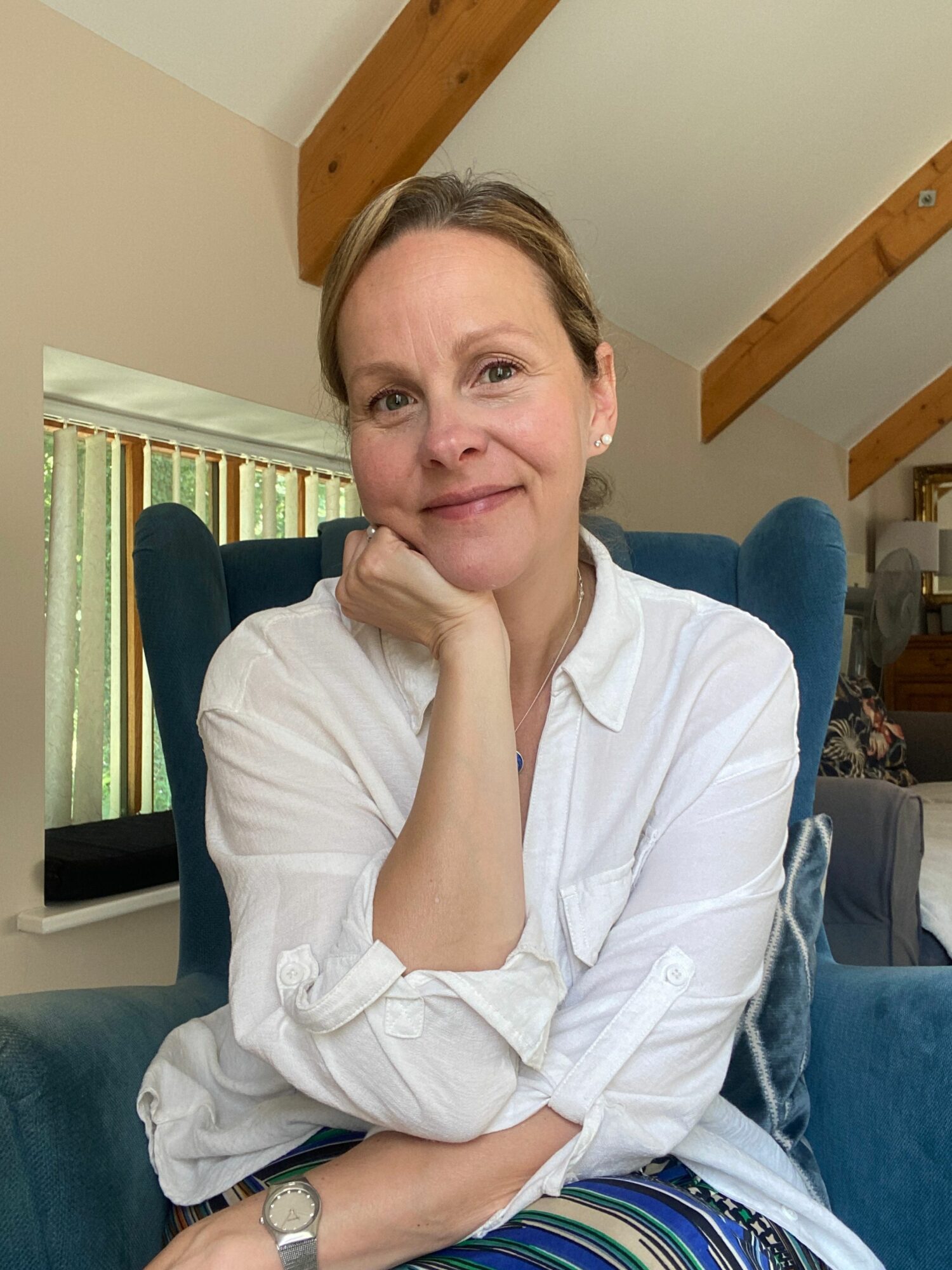 Woman with light brown hair resting her chin on her hand, sitting on a blue couch in a room with wooden beams.