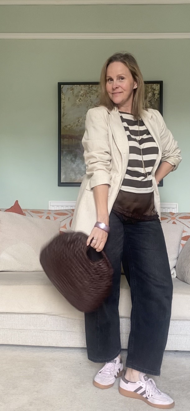 Woman standing in living room, holding a large dark bag, wearing striped shirt, beige jacket, jeans, and sneakers.