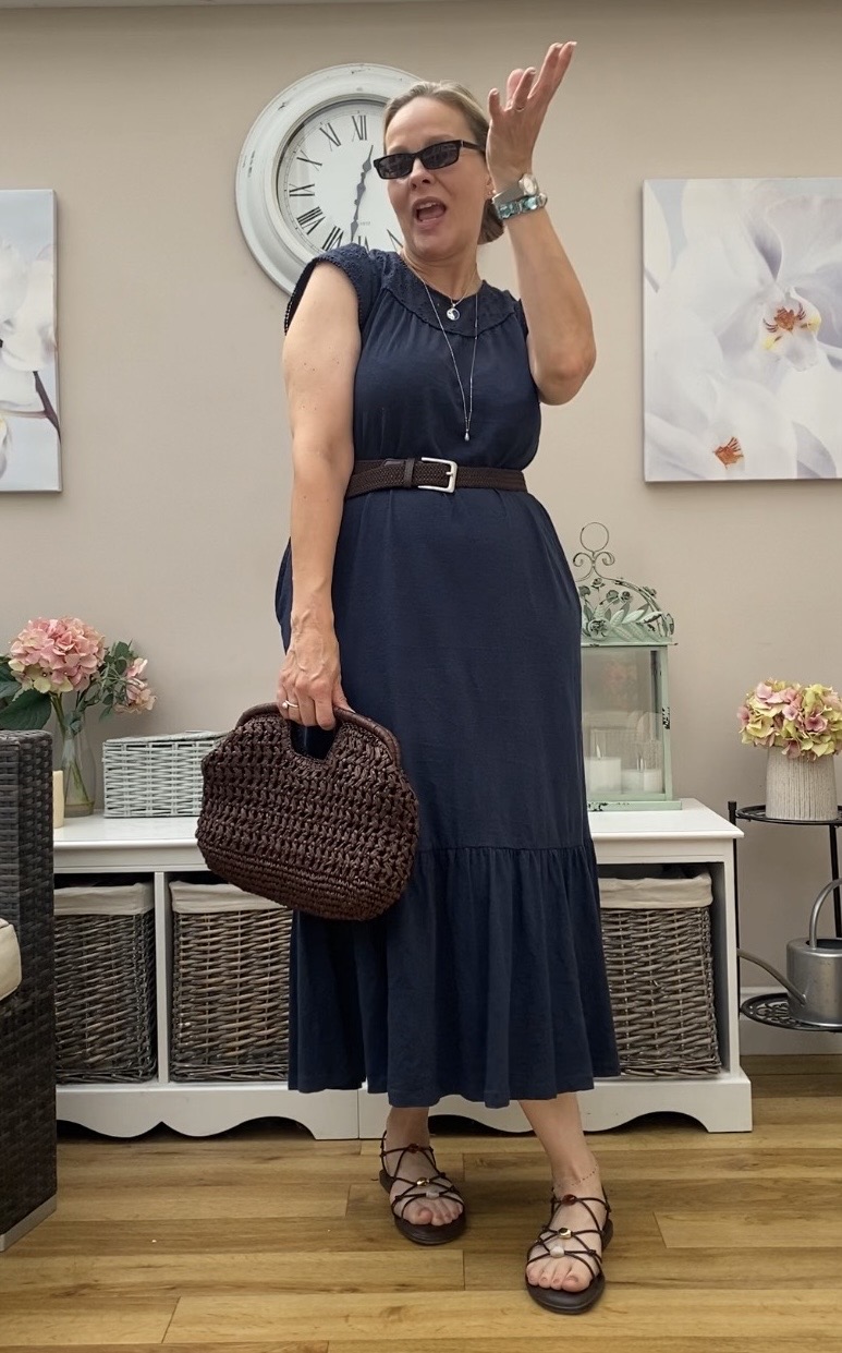 Woman in a navy dress with sunglasses, holding a woven bag, standing indoors with floral decorations and a clock behind her.
