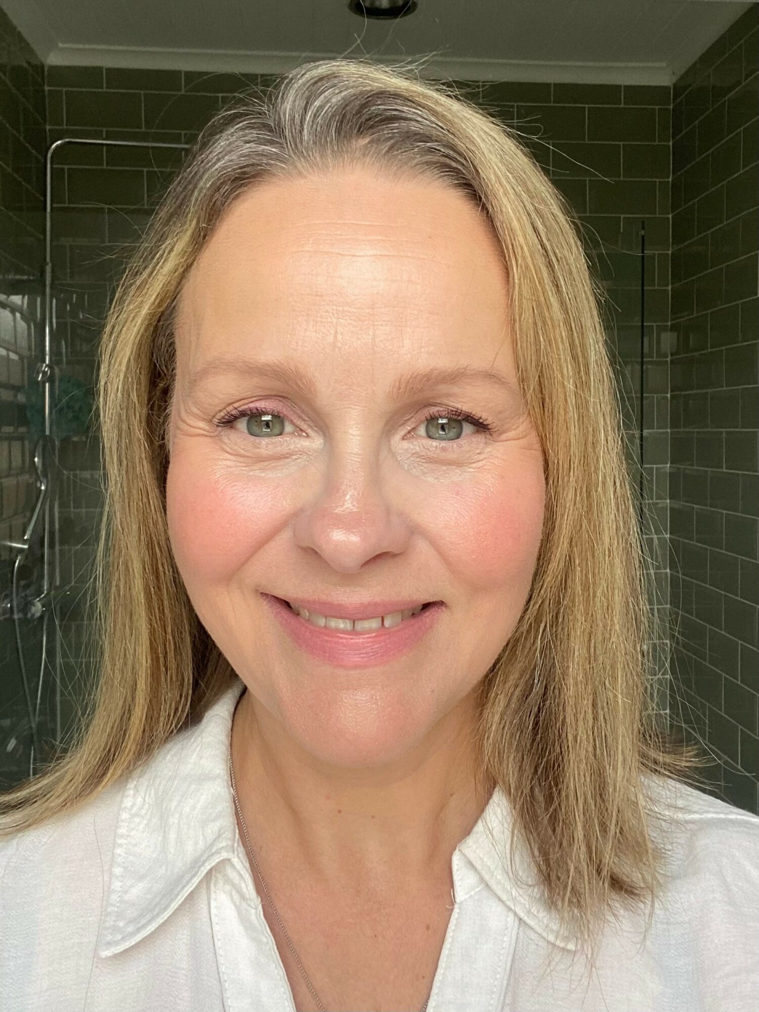 Close-up of a smiling woman with blonde hair and light-colored eyes, wearing a white shirt, in a tiled room.