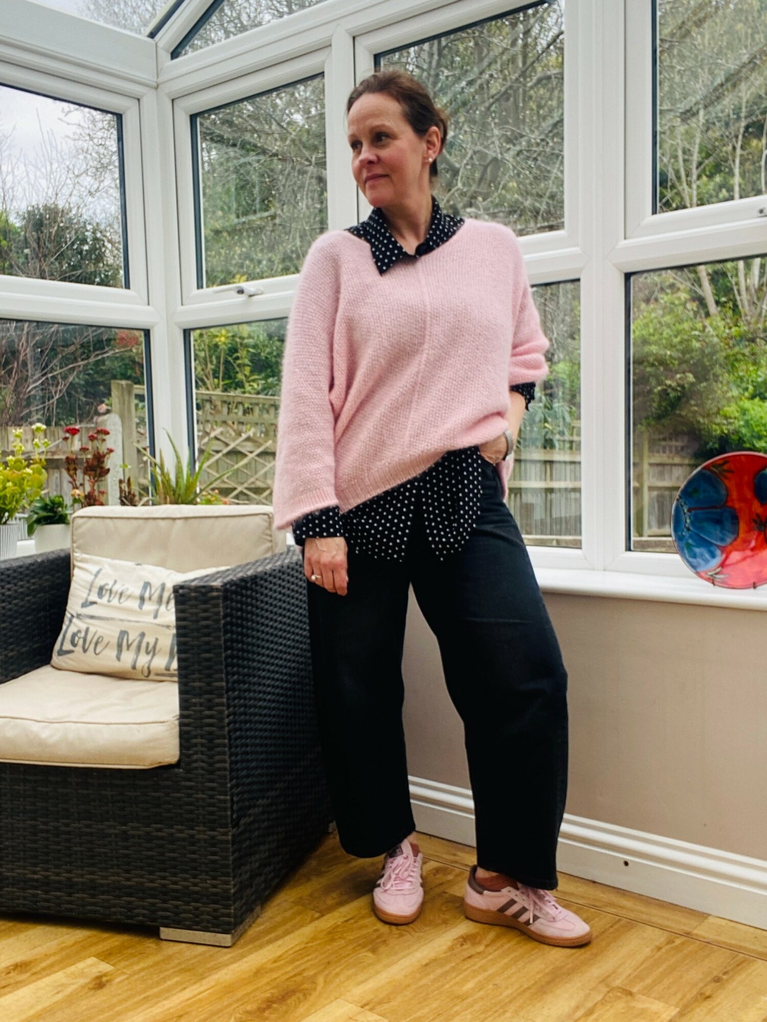 Woman standing indoors near windows, wearing pink sweater with black polka dot collar, black pants, and pink shoes.