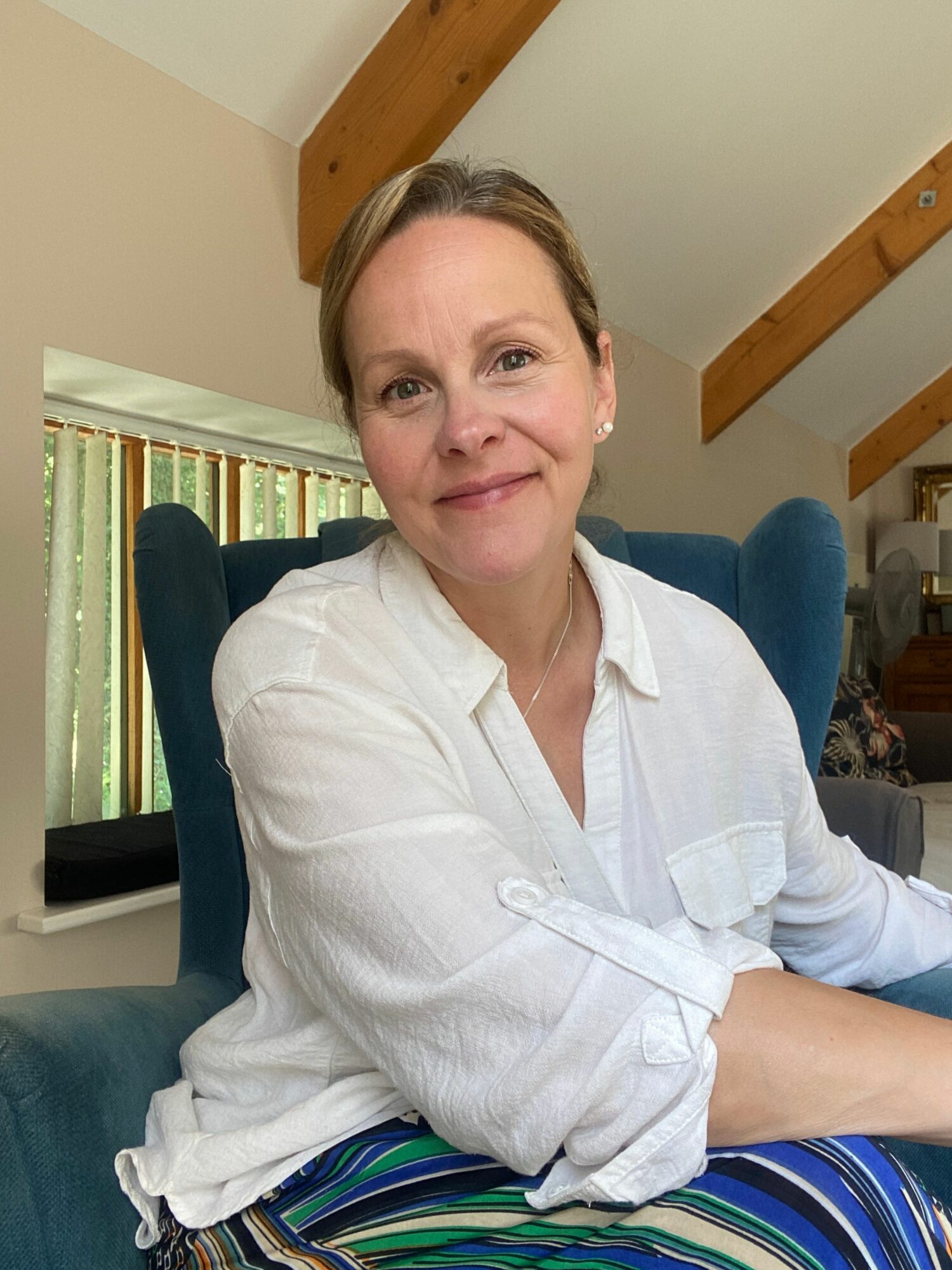 Woman sitting on a blue couch, wearing a white shirt and striped skirt, in a room with wooden beams and a railing.
