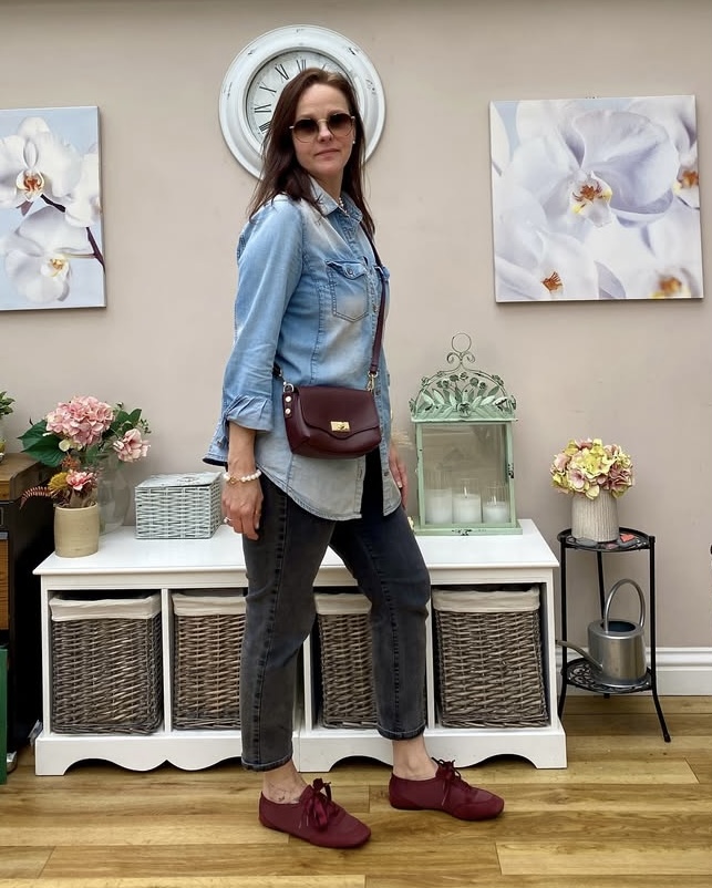 Woman with sunglasses standing in a room with floral artwork, a clock, and decorative items, wearing a denim jacket and burgundy shoes.