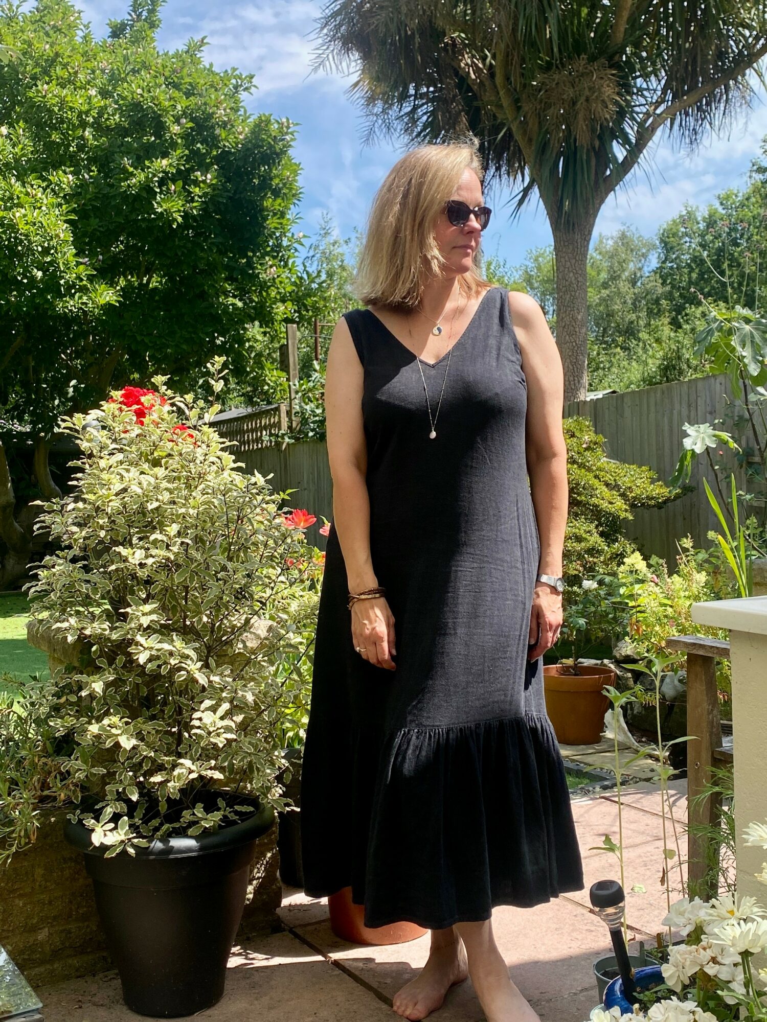 Woman in black dress and sunglasses standing outdoors in a garden with plants and trees.