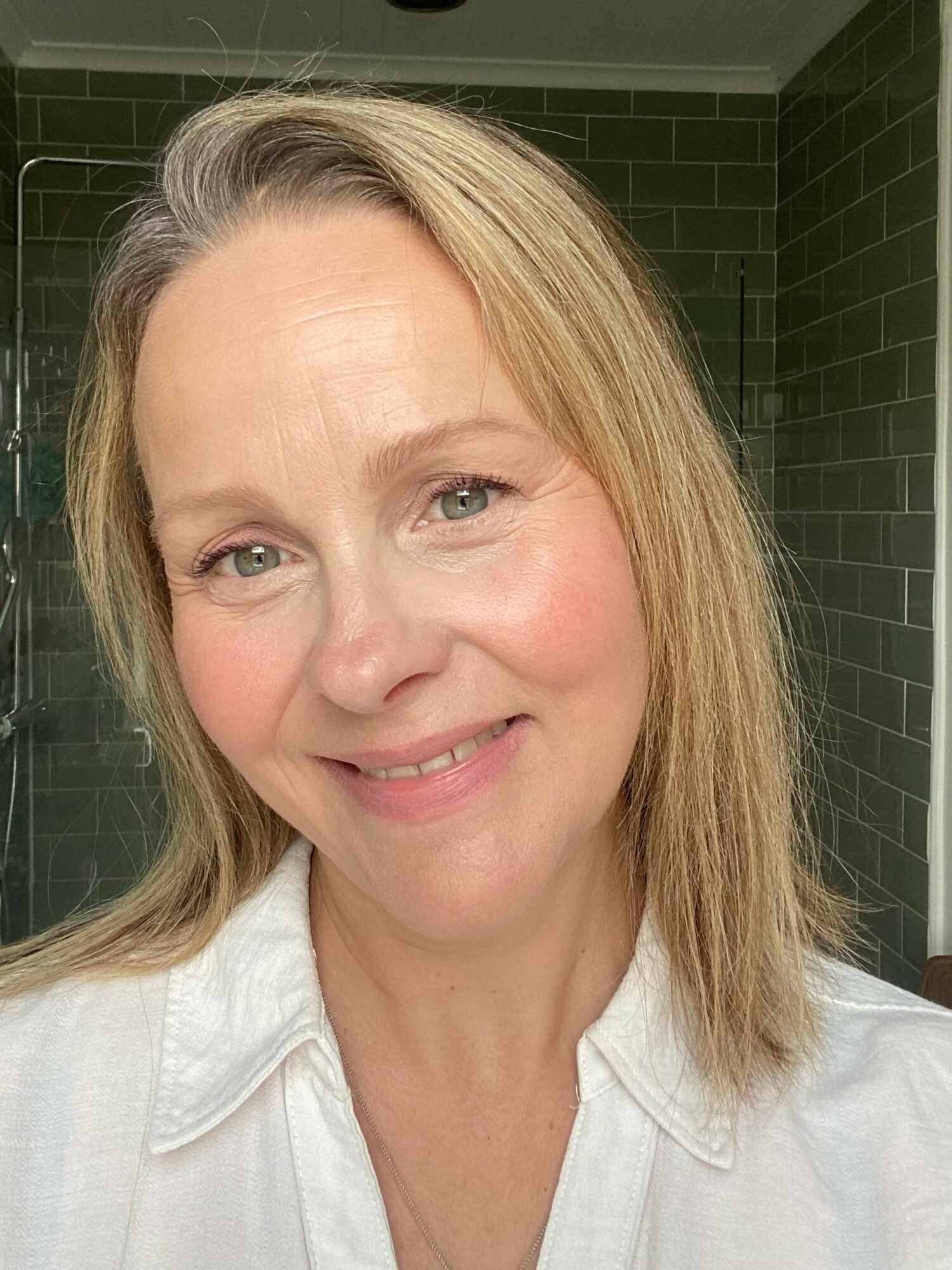 Close-up of a smiling woman with blonde hair and light skin, wearing a white shirt, in front of a green tiled wall.