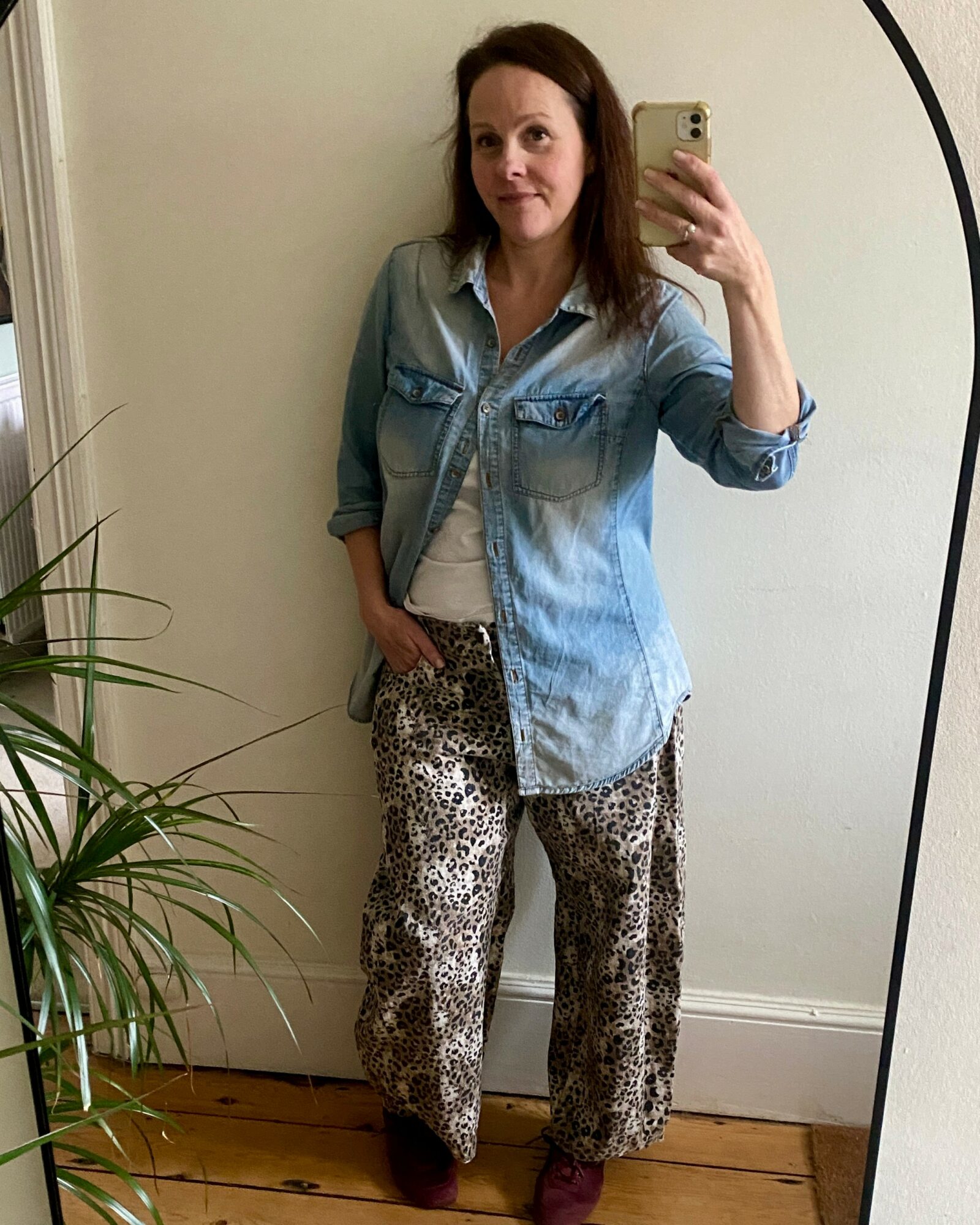 Woman taking a mirror selfie, wearing a denim shirt, white top, and leopard print pants, standing indoors near a plant.