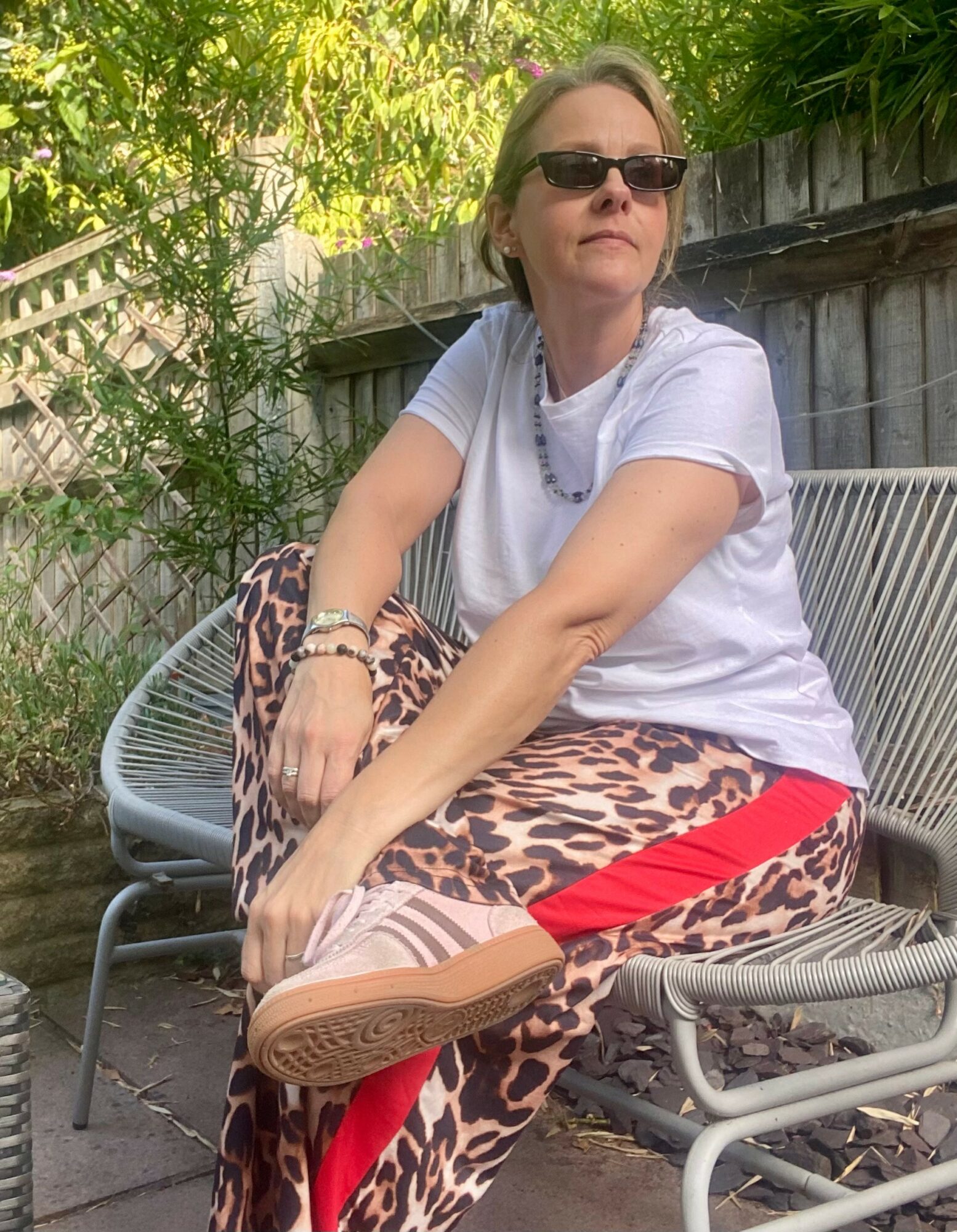 Woman sitting outdoors on a metal bench, wearing sunglasses, a white t-shirt, leopard print pants, and a red stripe, with greenery in background.