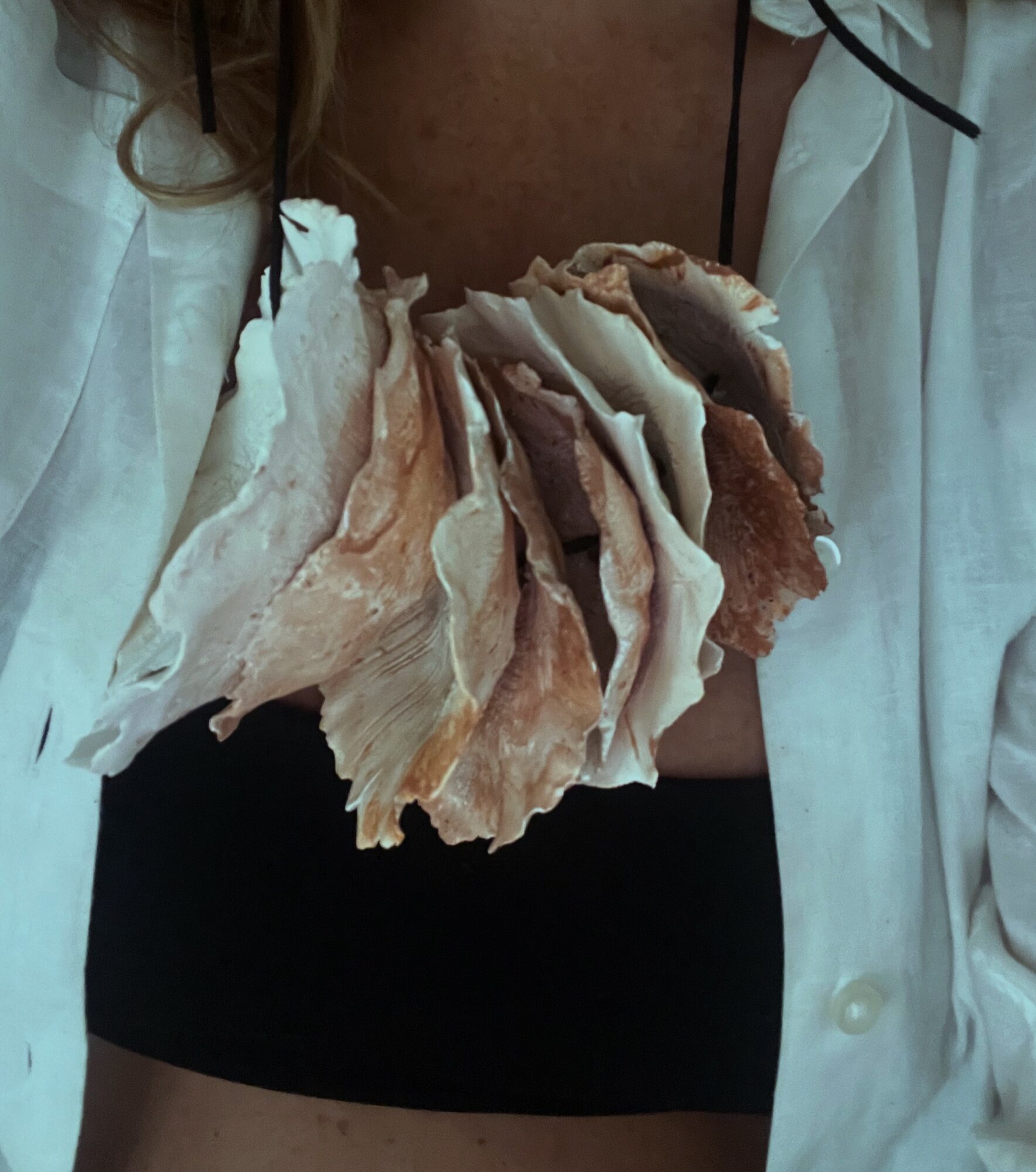Person wearing a white shirt with a large, layered, beige and brown shell necklace, black top visible underneath, close-up view.