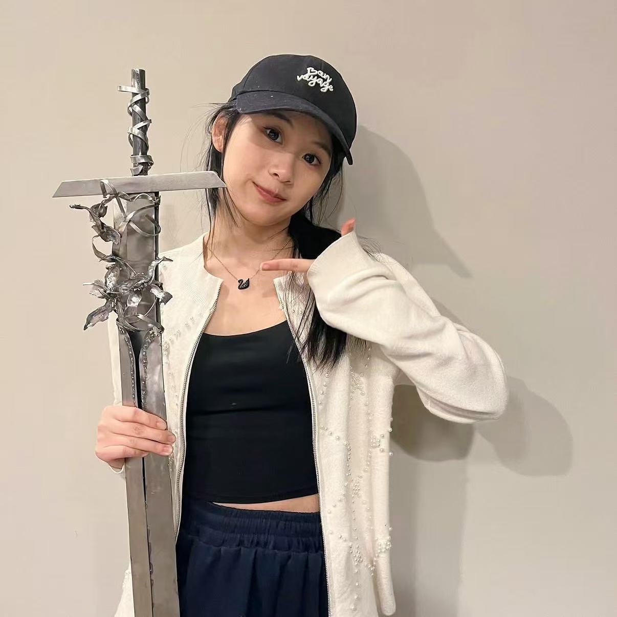 Young woman holding a sword, wearing a black cap, black top, and white jacket, standing against a plain wall.