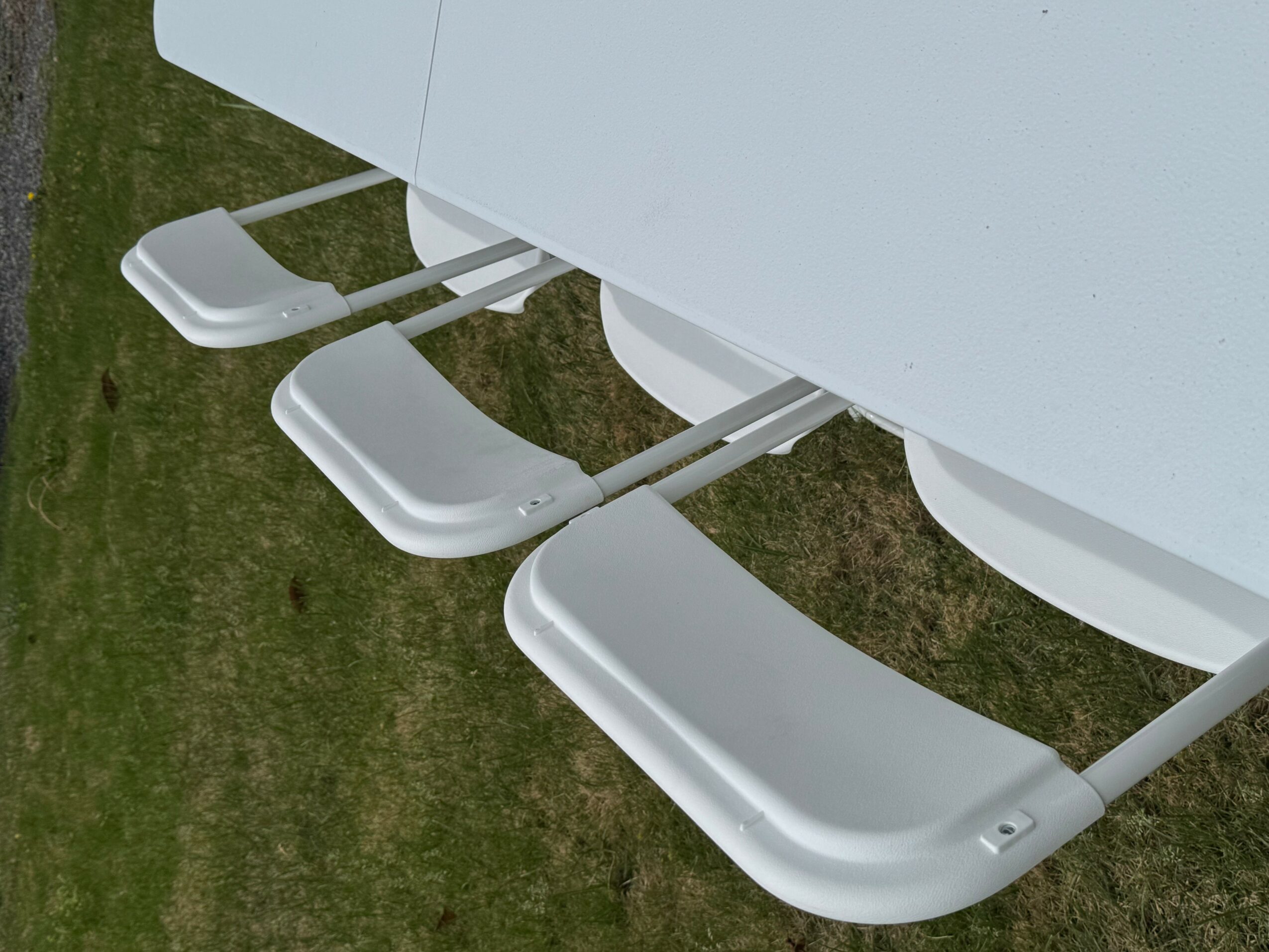 Four white chairs are stacked upside down on a white table outdoors, with grass visible below.