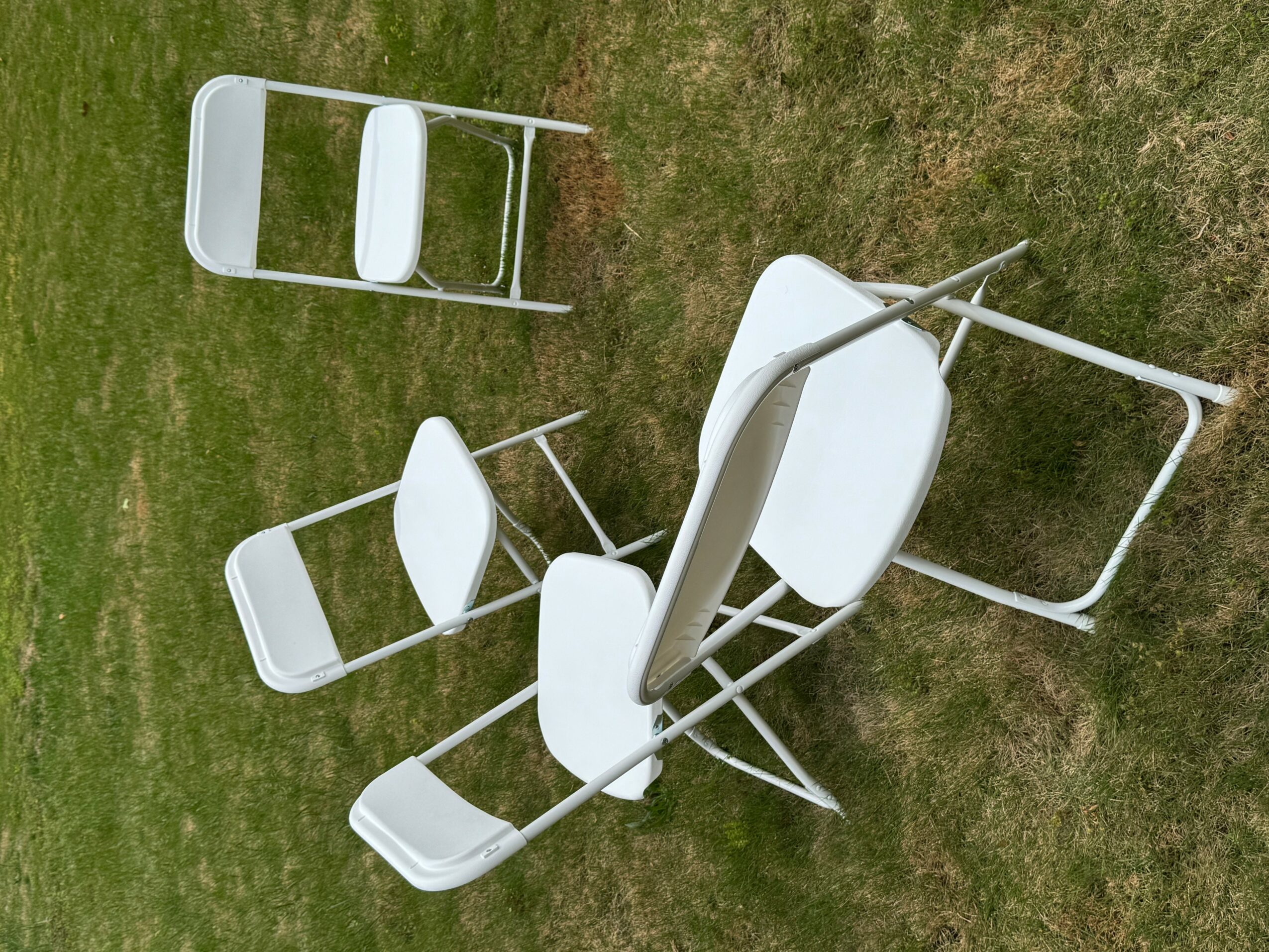 Several white chairs are stacked and fallen on grass, with some chairs leaning against each other and one upside down.