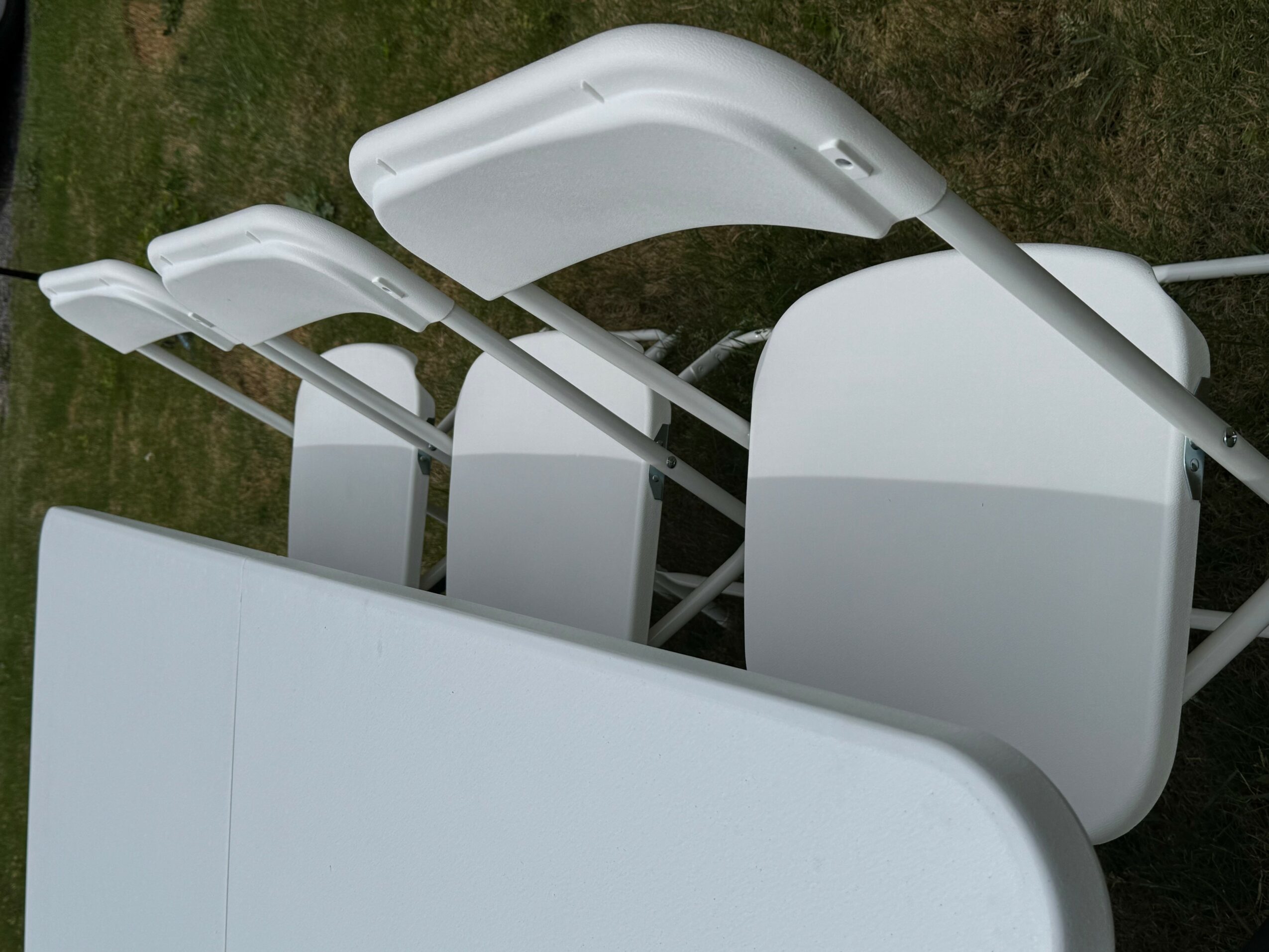 Three white outdoor lounge chairs on grass, viewed from the side, with curved armrests and backrests.
