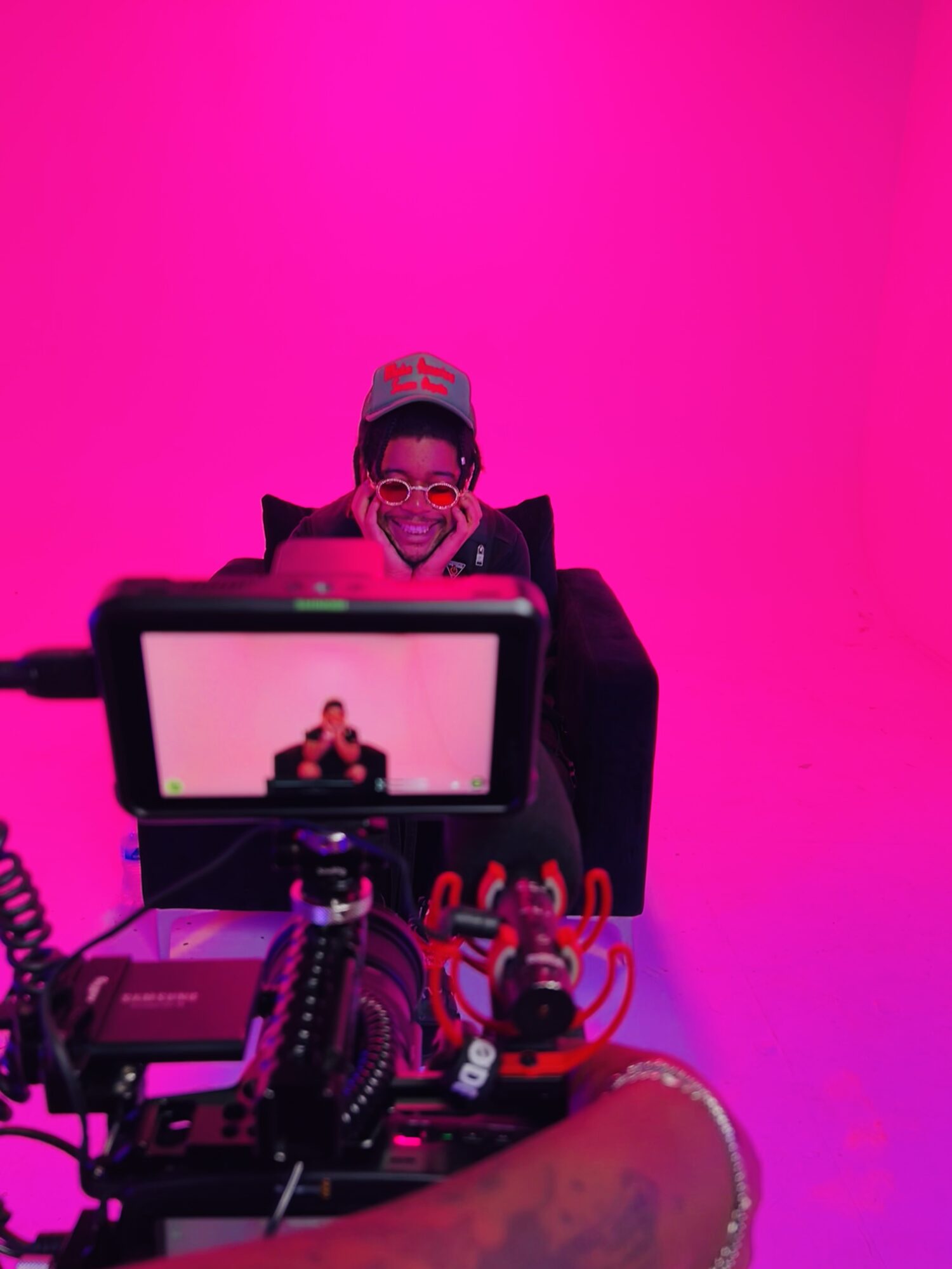 Person wearing glasses and a mask, sitting behind a camera setup, against a pink background.