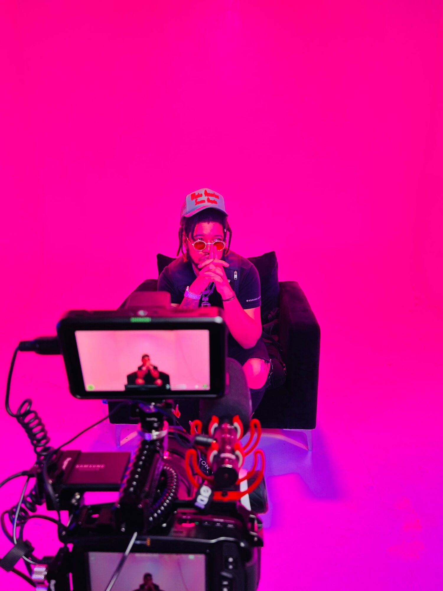 Person sitting on a black chair with a pink background, wearing glasses and a cap, surrounded by camera equipment and a toy figure.