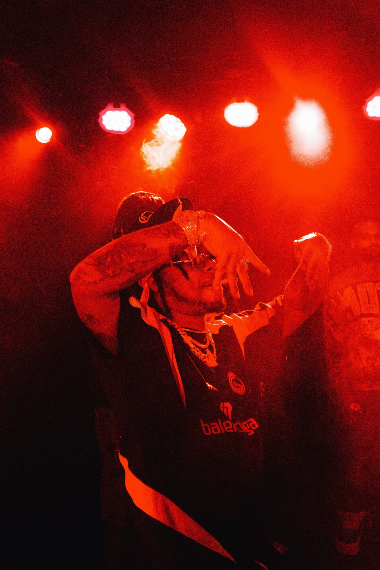 Person performing on stage with red lighting, holding a microphone, wearing dark clothing, and a cap.