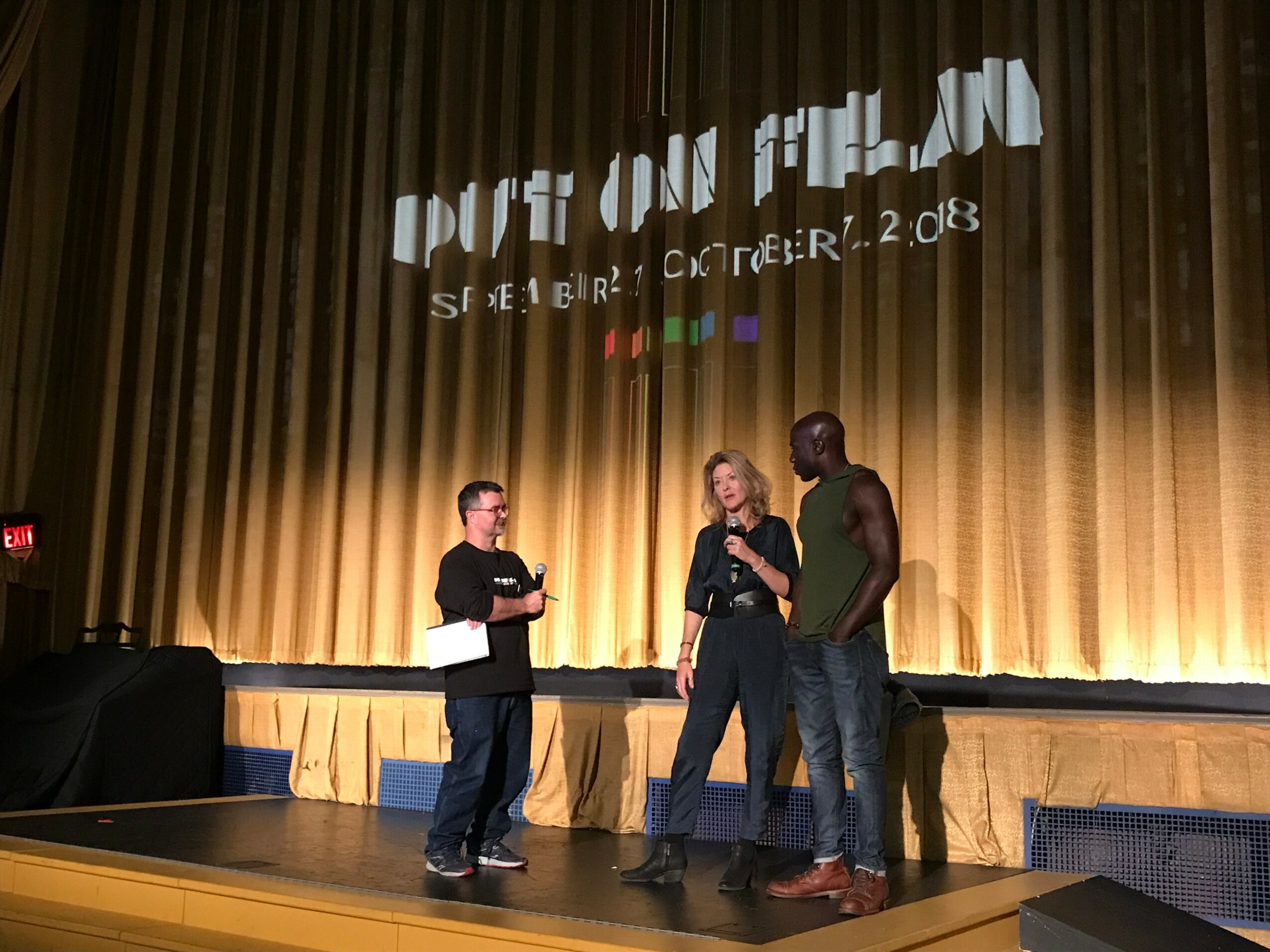 Three people stand on a stage with a gold curtain, engaging in conversation, with a projected event logo above them.