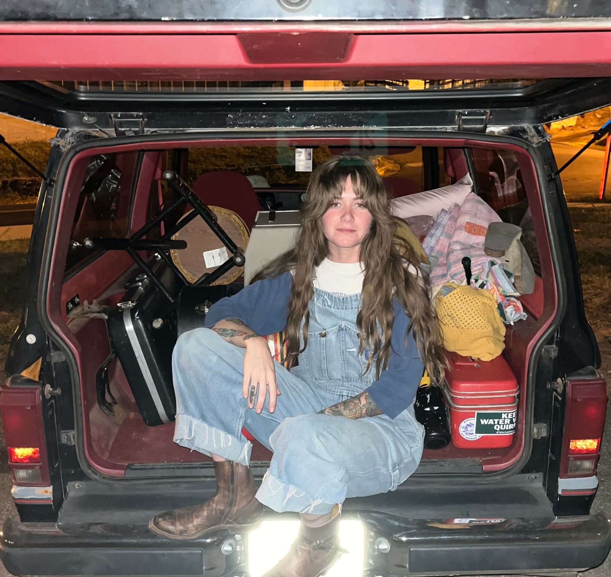 Person with long hair sitting in the open trunk of a vehicle, surrounded by bags and items, wearing denim overalls and boots.