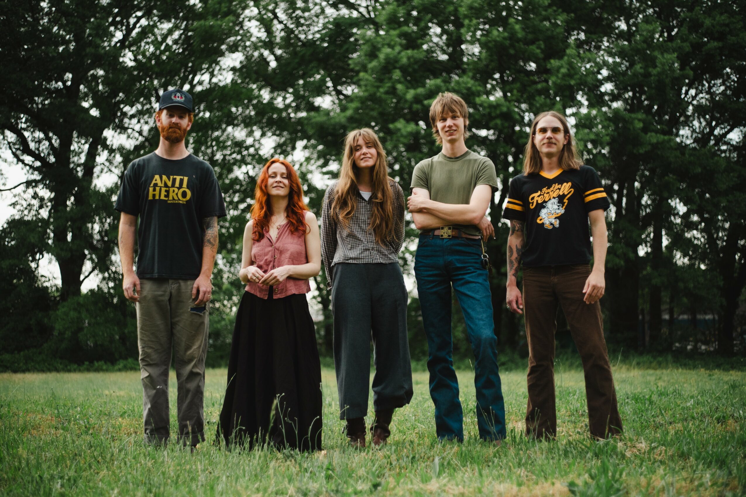Five people standing outdoors on grass with trees in the background, facing forward, in casual clothing.