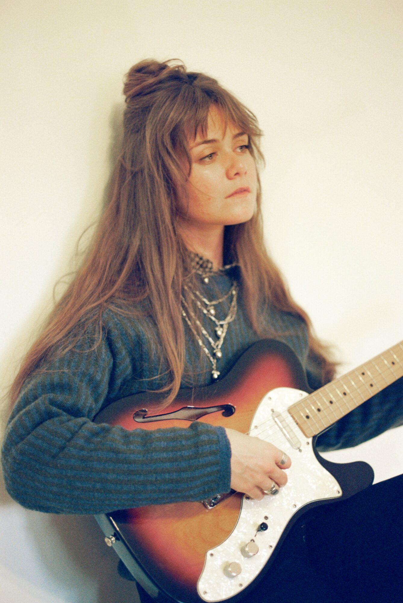 Young woman with long hair and layered hairstyle, wearing a dark sweater and multiple necklaces, holding an electric guitar.