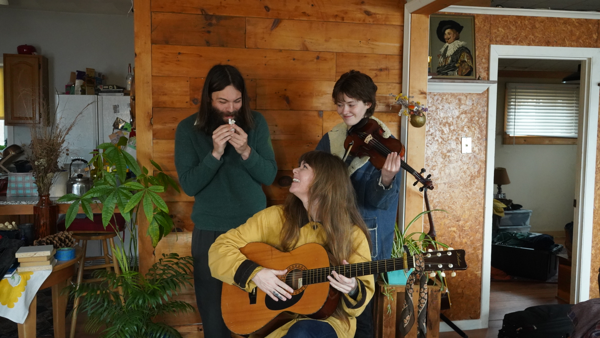 Three people in a cozy room, one playing guitar, two singing, with plants and wooden walls visible.