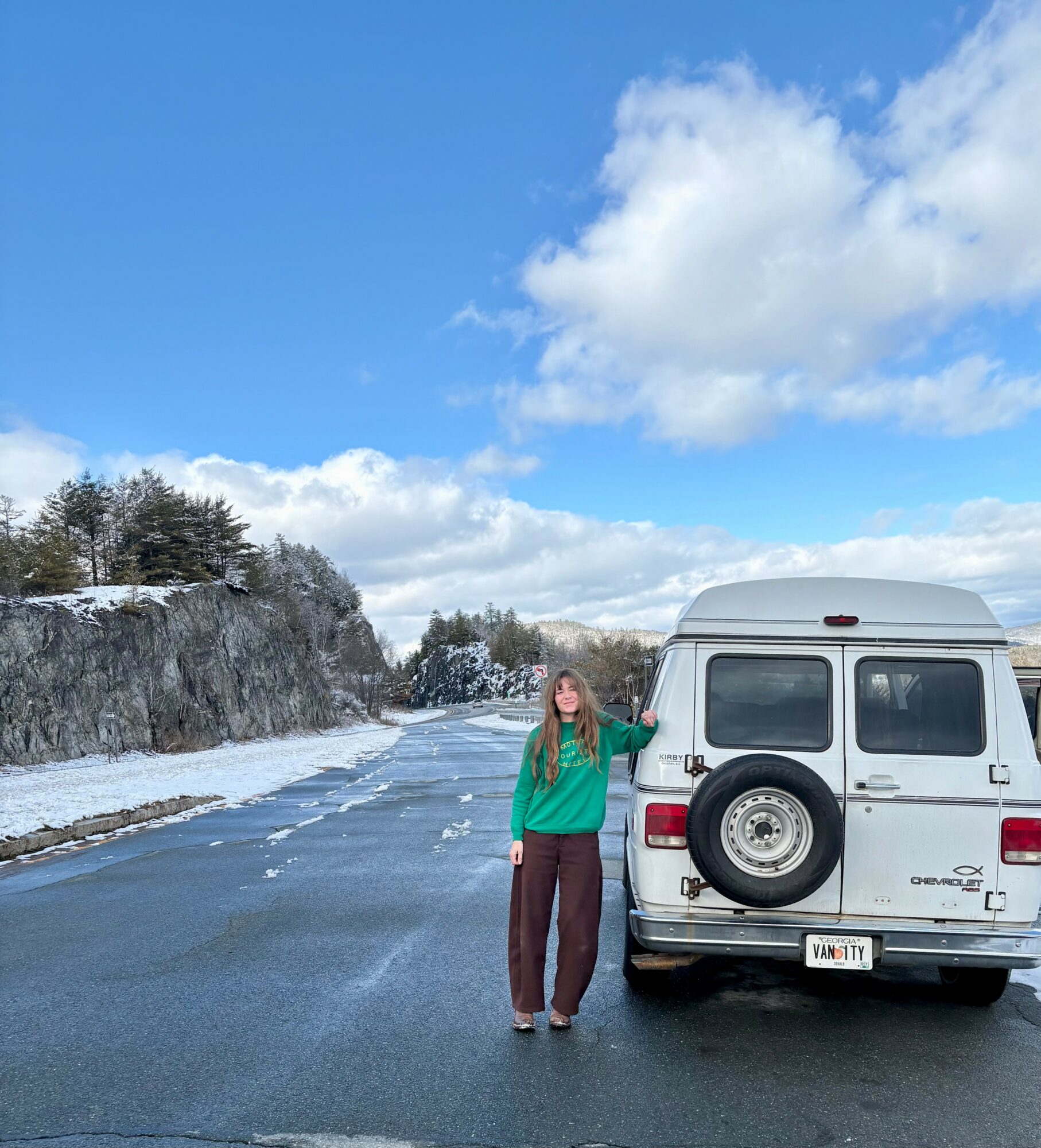 Person standing next to a white van on a snowy roadside with a cliff and trees in the background, under a partly cloudy sky.