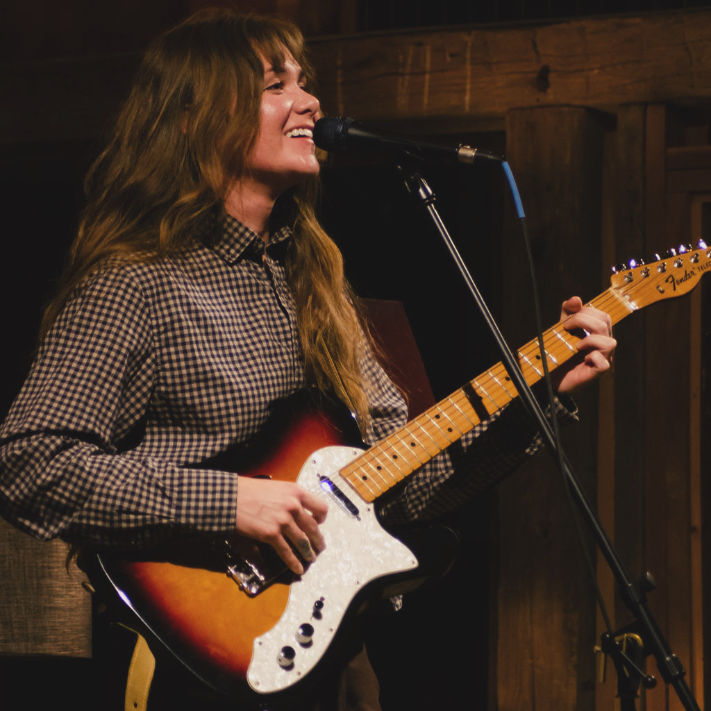Woman playing electric guitar and singing into microphone on stage, smiling, with wooden background.