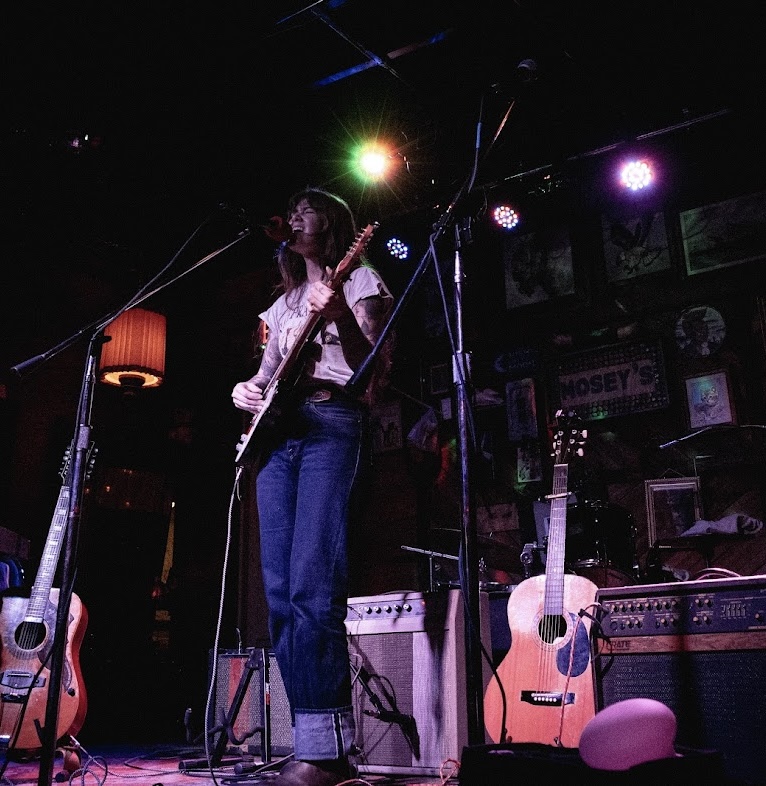 Person playing guitar on stage with colorful lights, surrounded by musical equipment and posters, in a dimly lit venue.