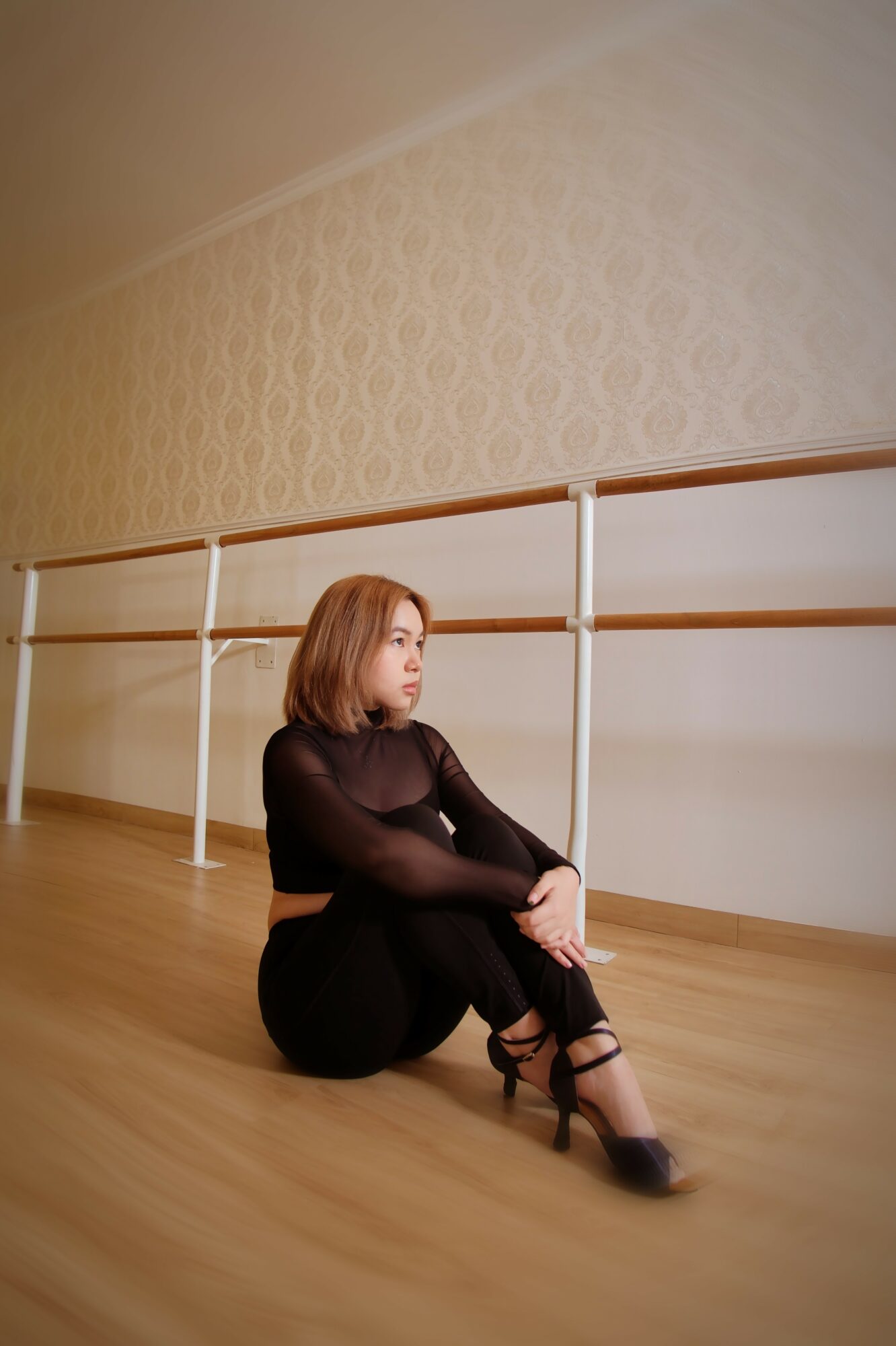 Woman sitting on the floor with crossed legs, wearing black clothing and high heels, in a dance studio with wooden floor and ballet barre.