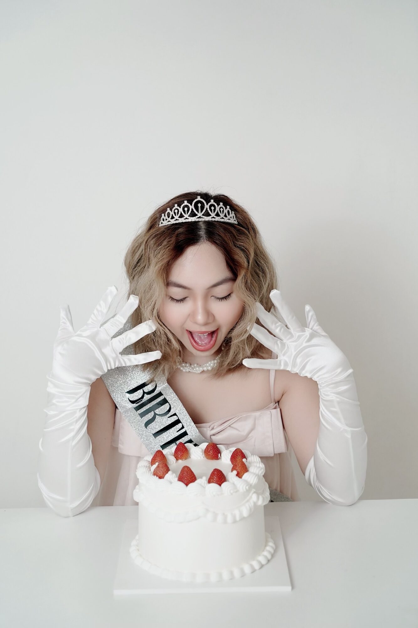 Young woman wearing a tiara and sash celebrating birthday with a cake topped with strawberries, smiling with hands raised.